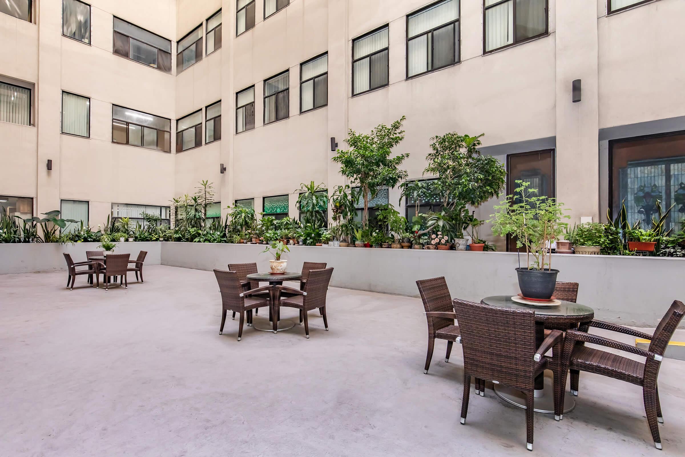 A spacious courtyard featuring several tables and chairs made of wicker. The area is filled with greenery, including potted plants and small trees, surrounded by tall building walls with large windows. The atmosphere is calm and inviting, perfect for relaxation or outdoor gatherings.