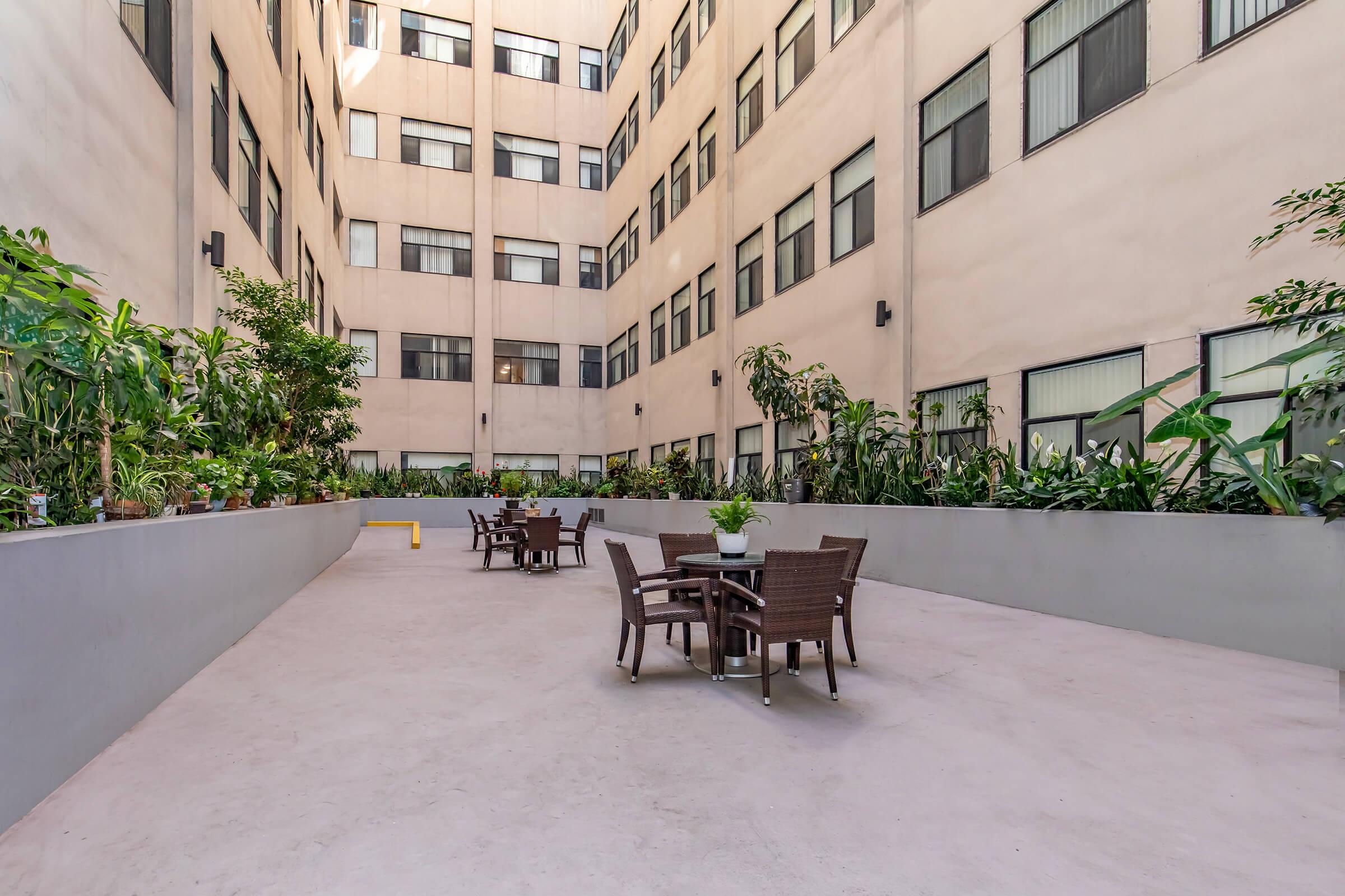 A spacious courtyard surrounded by buildings, featuring several potted plants and trees. In the center, there are outdoor tables and chairs arranged for seating, providing a relaxing outdoor space in an urban setting. Natural light filters through the surrounding windows.