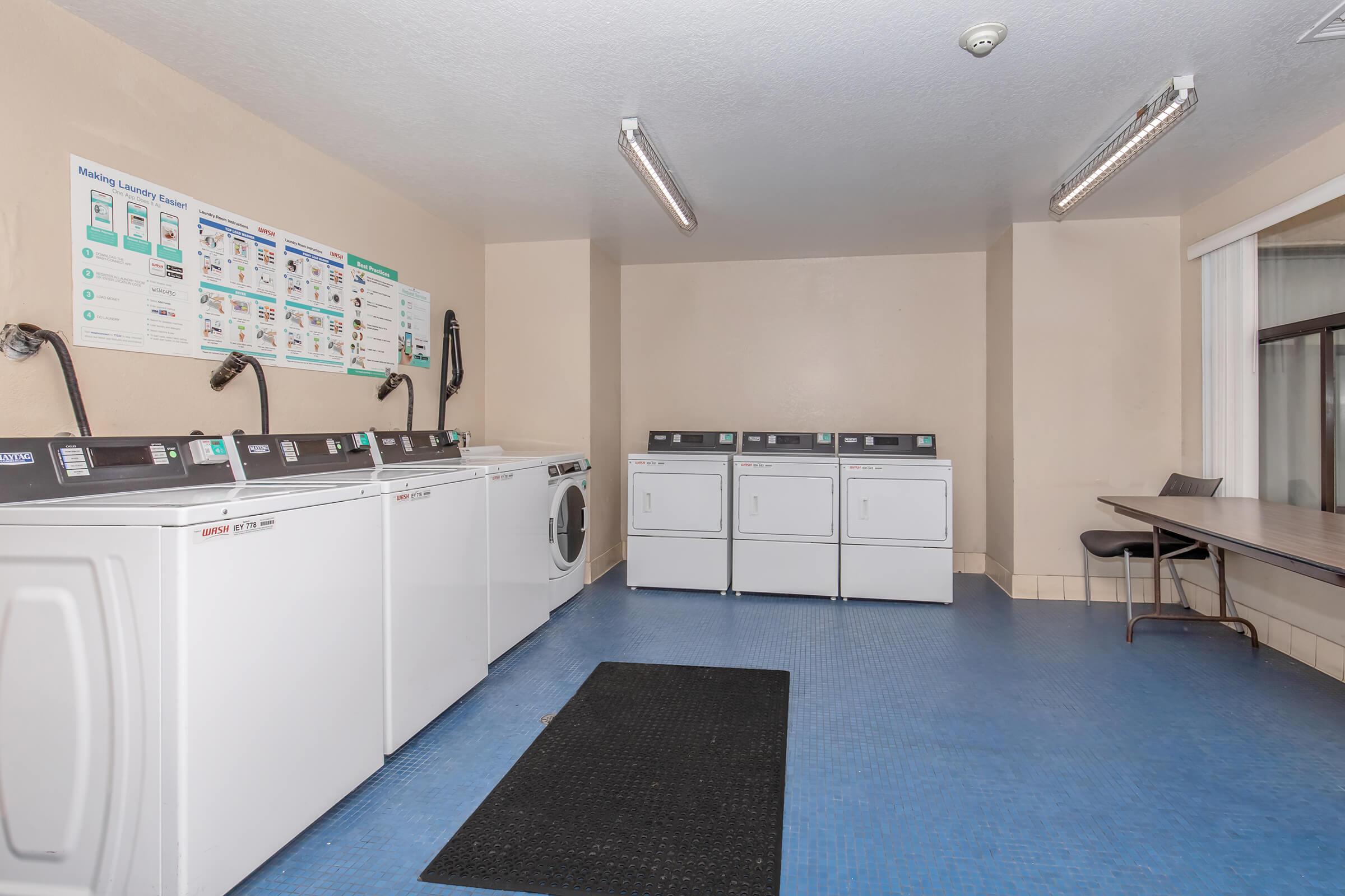 A clean and well-lit laundry room featuring several washers and dryers arranged in a row against the wall. A large informational poster is mounted above the machines. There is a table and chair to the side, and the floor is covered with blue vinyl. The space is tidy and organized, suitable for laundry activities.