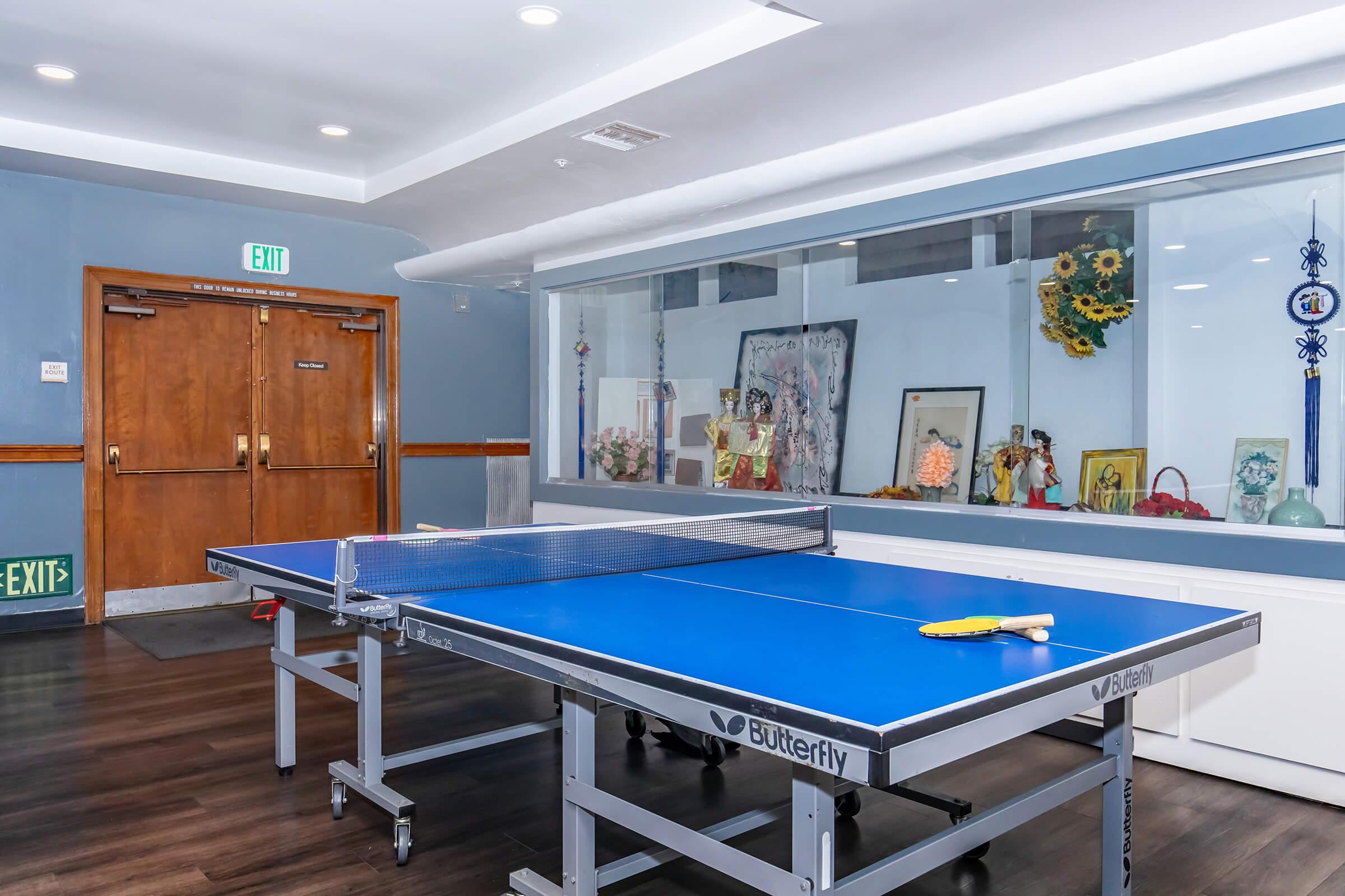A blue table tennis table with paddles positioned on it, set in a room featuring wooden double doors in the background. The walls are adorned with framed art and decorative items, including flowers, creating a vibrant environment for recreational activities. Exits are marked with green signs.