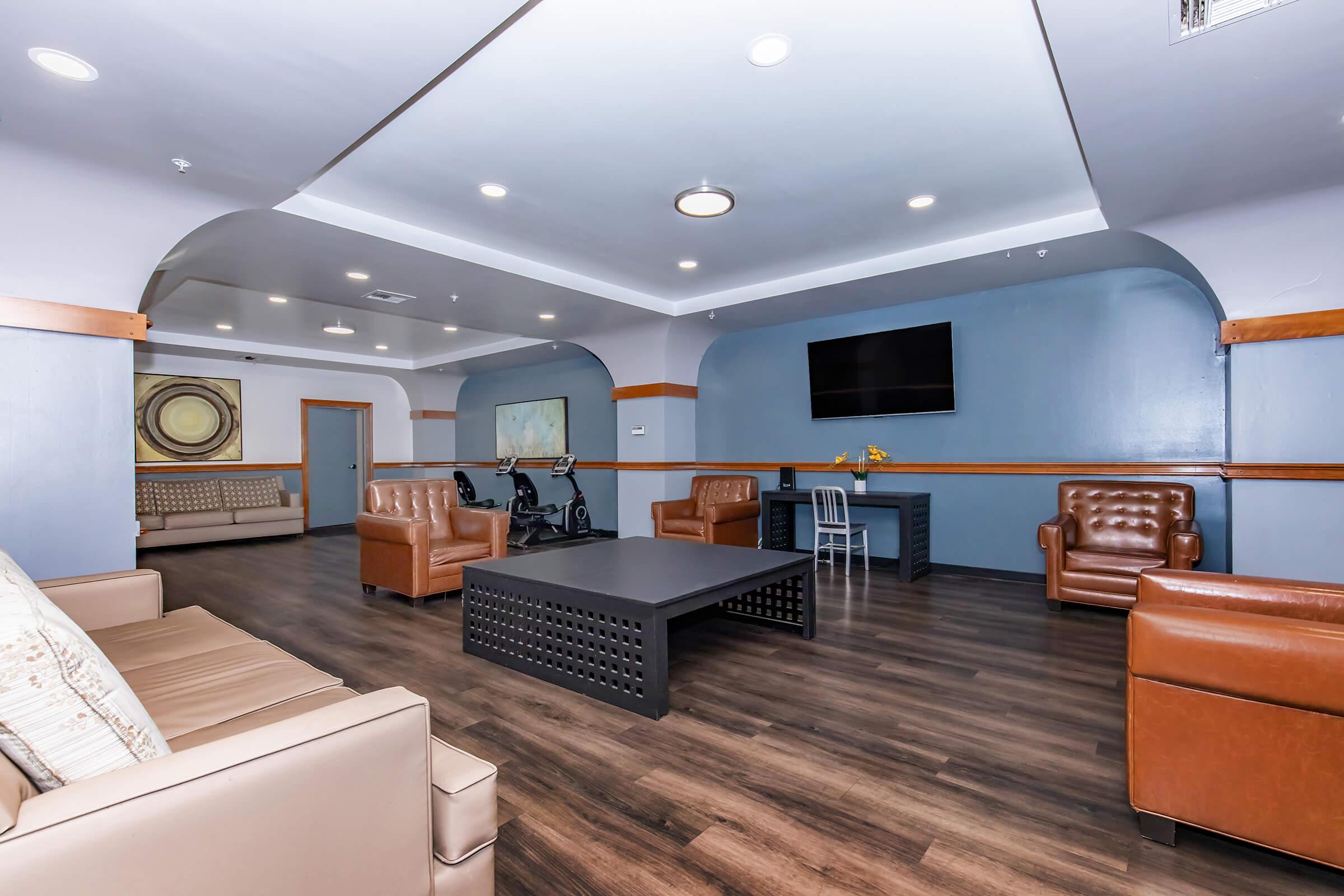 A modern lounge area featuring a comfortable seating arrangement with leather chairs and a sofa, a large black coffee table, a wall-mounted TV, and a small desk. The room has a sleek design with wooden accents and neutral colors, creating a welcoming environment for relaxation or socializing.