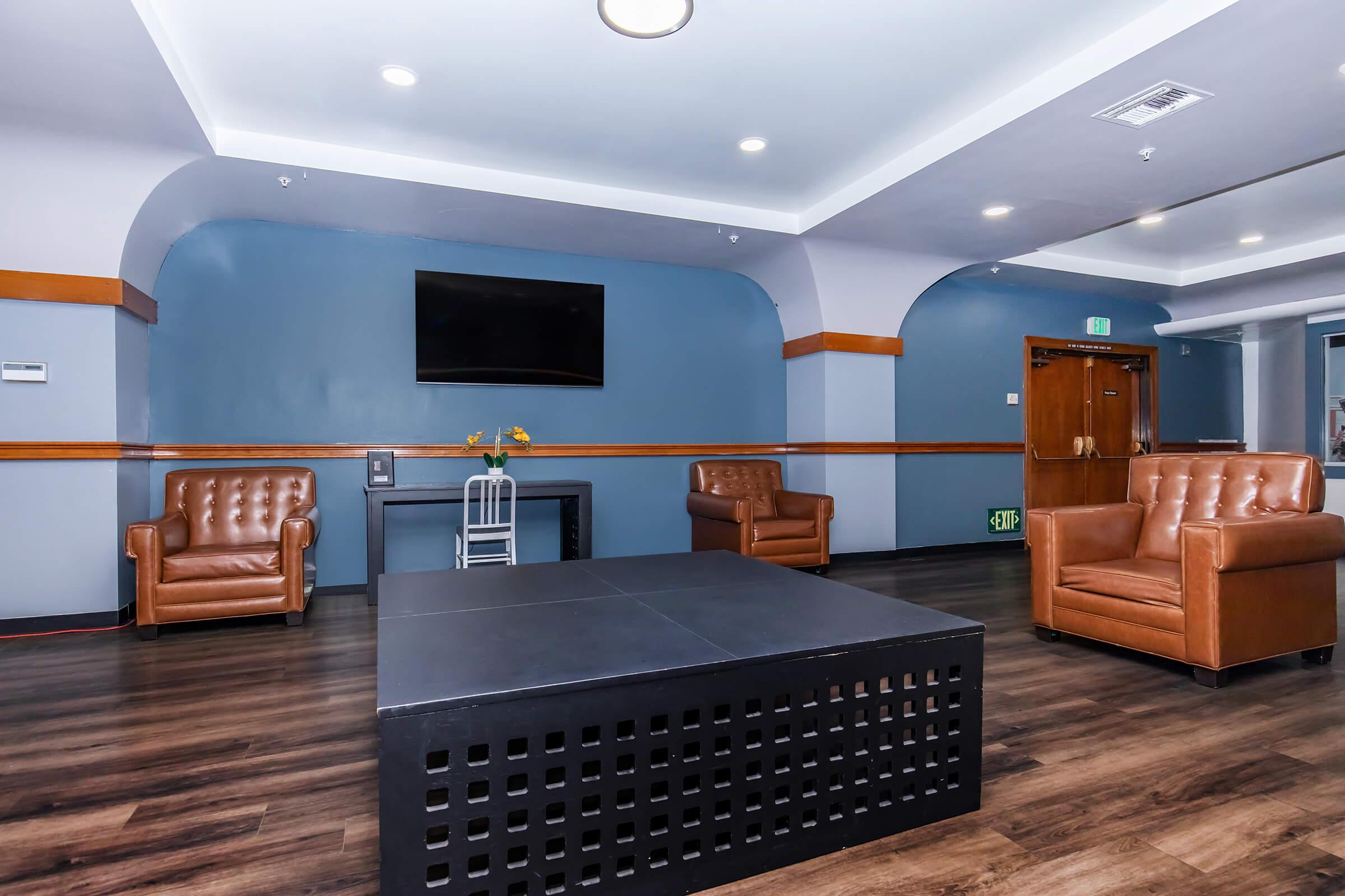A modern lounge area featuring two brown leather armchairs and a low black coffee table with a grid design. There is a flat-screen TV mounted on a blue wall, with a small decorative vase on a side table. The flooring is dark wood, and the room has a contemporary atmosphere.
