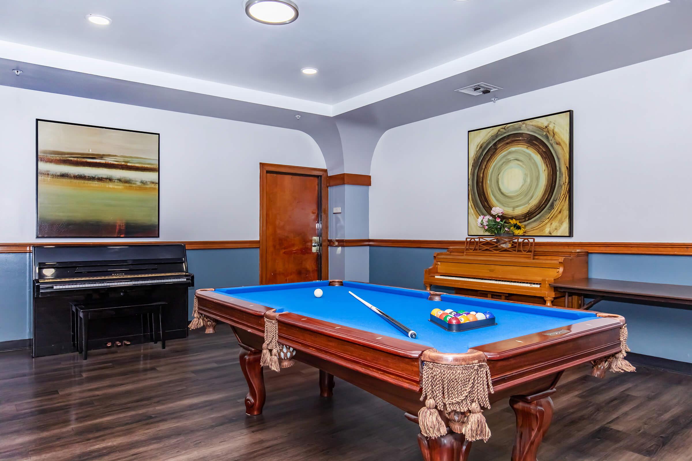 A room featuring a blue felt pool table with a cue and balls, beside a black piano and a wooden piano. The walls are decorated with two abstract paintings, and the floor has a dark wood finish. Soft lighting illuminates the space, giving it a cozy atmosphere.