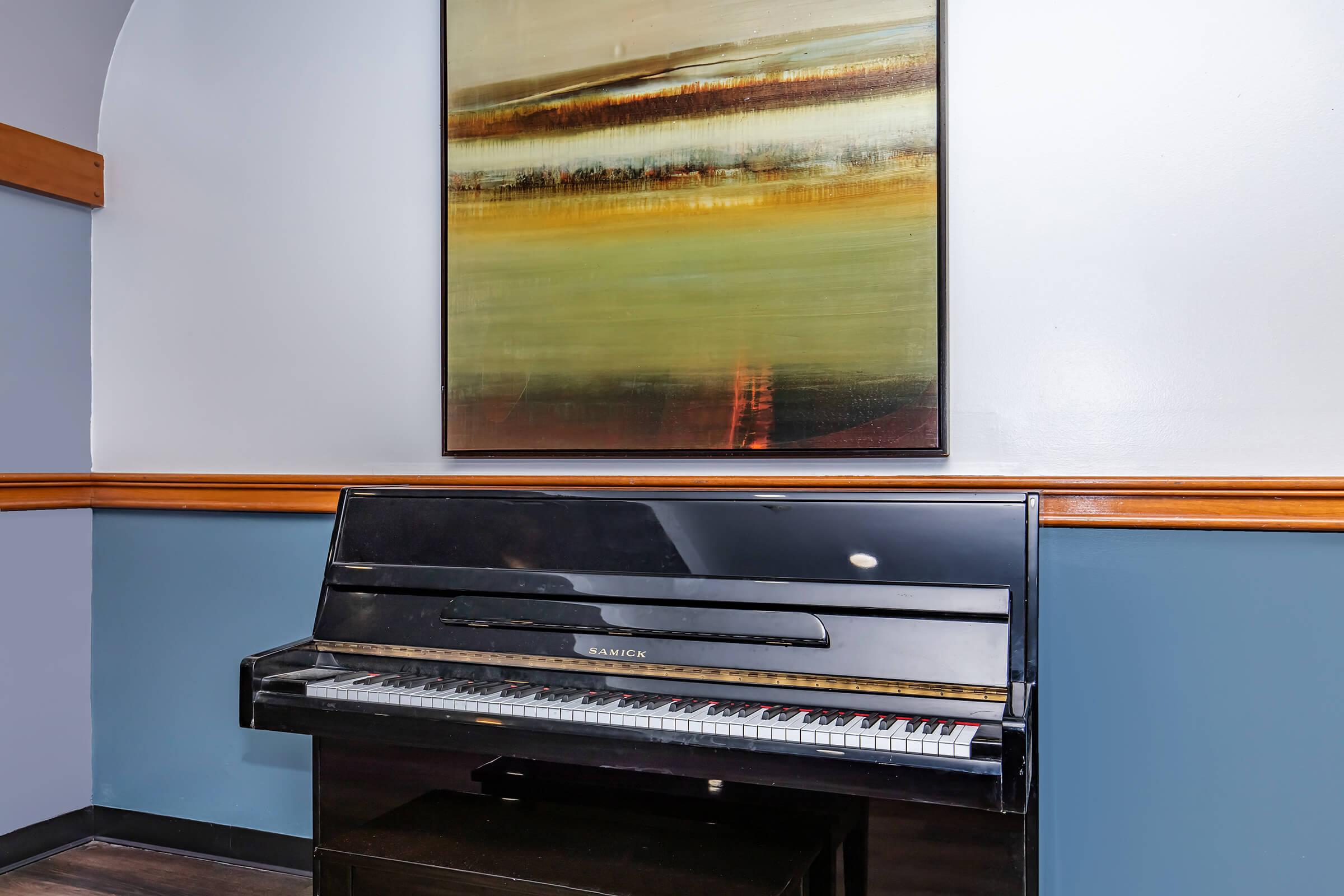 A black piano is positioned against a wall with blue and beige accents. Above the piano hangs a large abstract painting featuring earthy tones and horizontal stripes. The setting suggests a stylish and serene atmosphere, ideal for music and creativity.