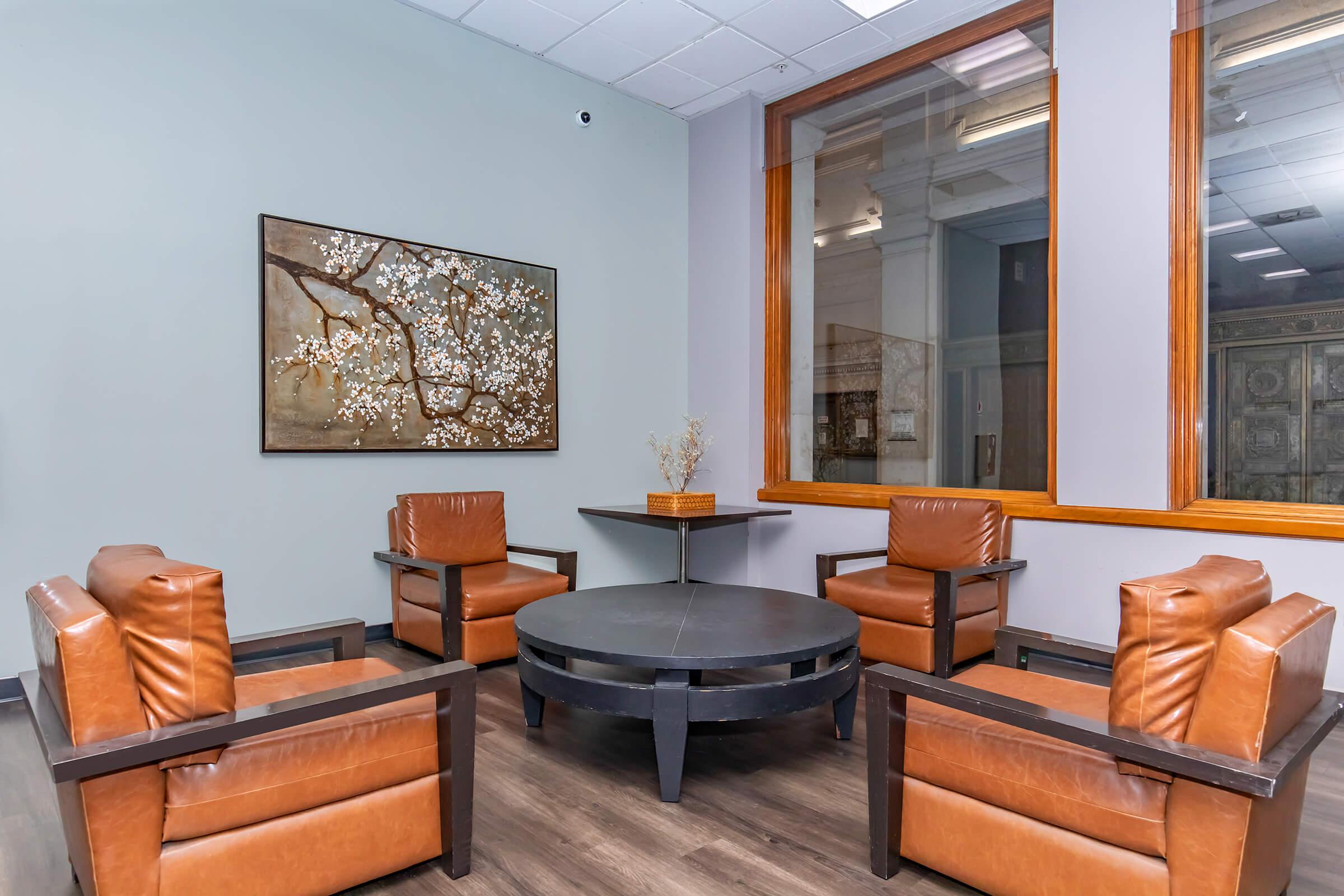 A modern sitting area featuring four brown leather armchairs arranged around a circular black table. A decorative wall art piece with a floral design hangs on the gray wall. Large windows provide natural light, and the flooring is a dark wood finish.