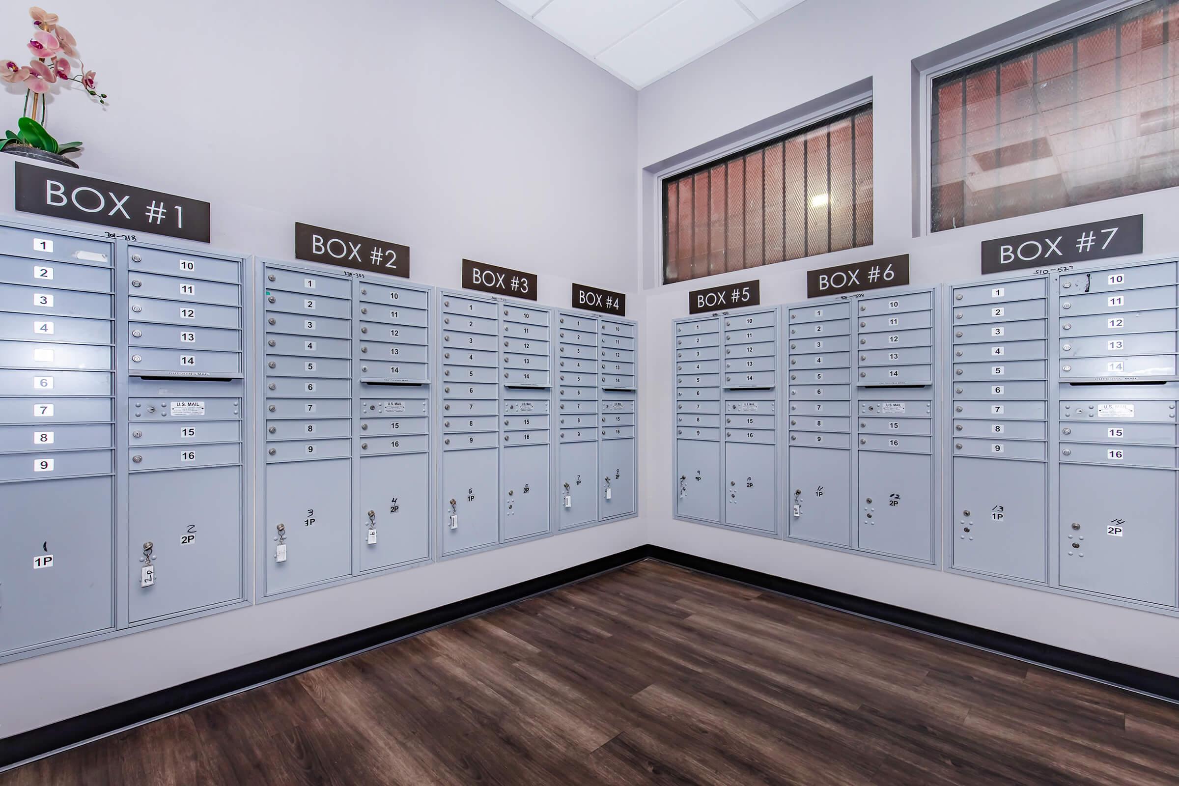 A well-lit room featuring multiple grey mailboxes arranged on two walls. Each mailbox is numbered, ranging from Box #1 to Box #7, with individual compartments for mail. Large windows allow natural light to enter, enhancing the modern and organized appearance of the space. The floor is dark wood.