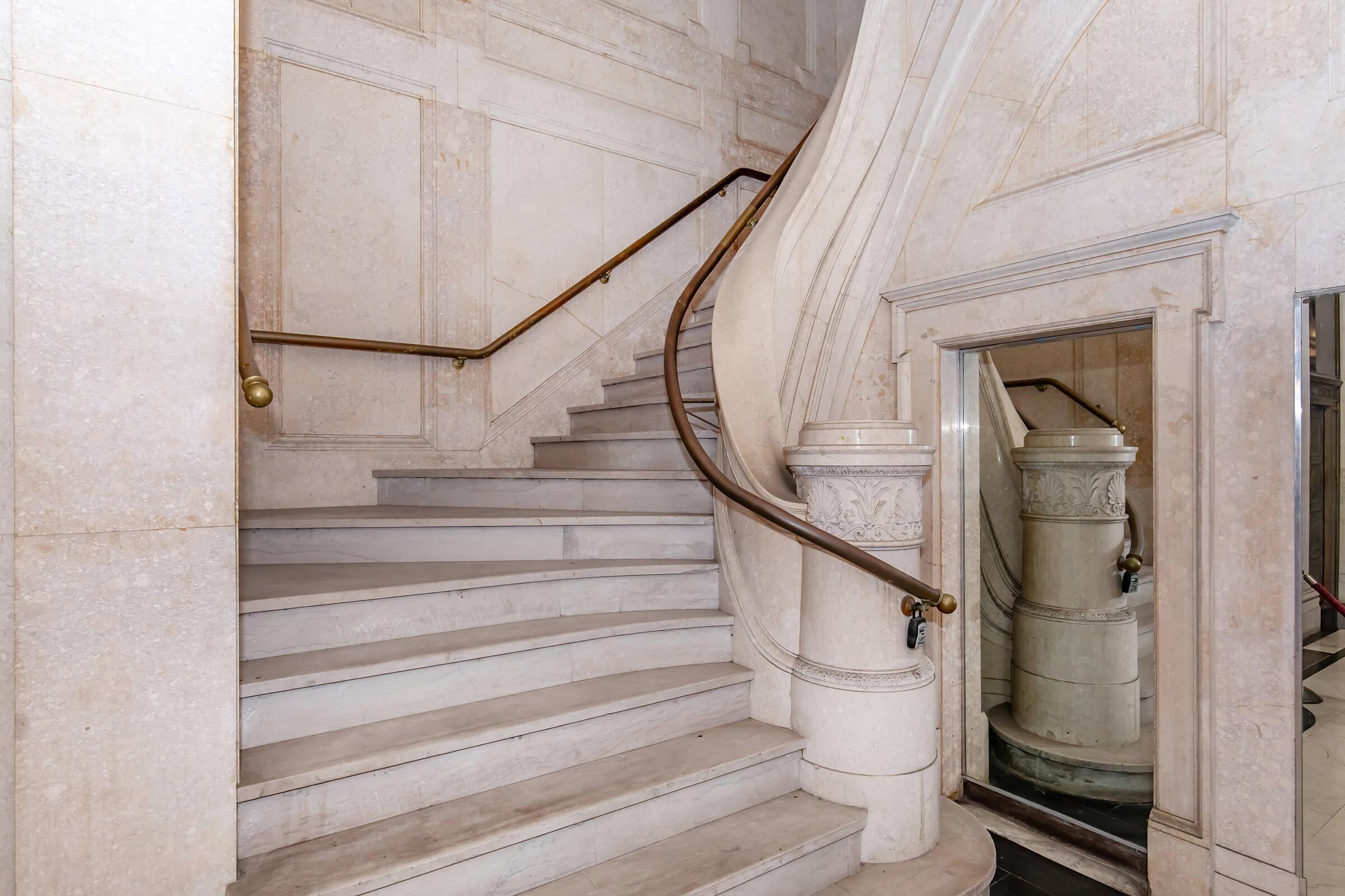 A curved staircase made of light-colored stone, featuring a decorative bannister. The staircase ascends with a classic design, and a mirrored wall reflects part of the staircase. The overall ambiance conveys elegance and architectural detail in a well-lit interior space.