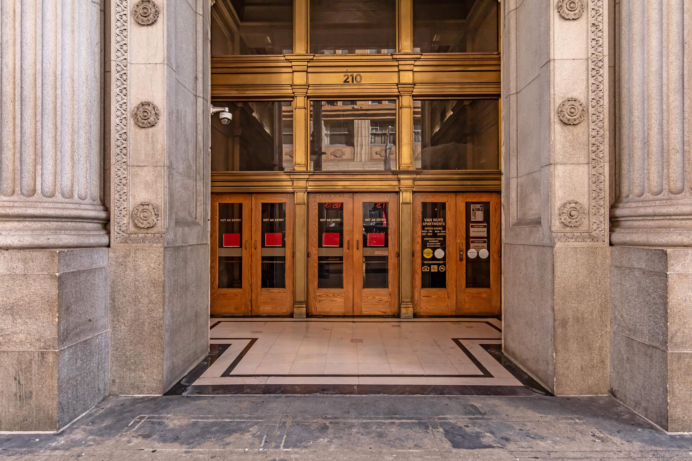 Entrance of a historic building featuring large wooden doors with ornate brass fixtures, flanked by columns. The floor is tiled in a decorative pattern. Red and white signs are displayed on the glass doors, indicating entry instructions or restrictions. The overall architecture showcases classic detailing.