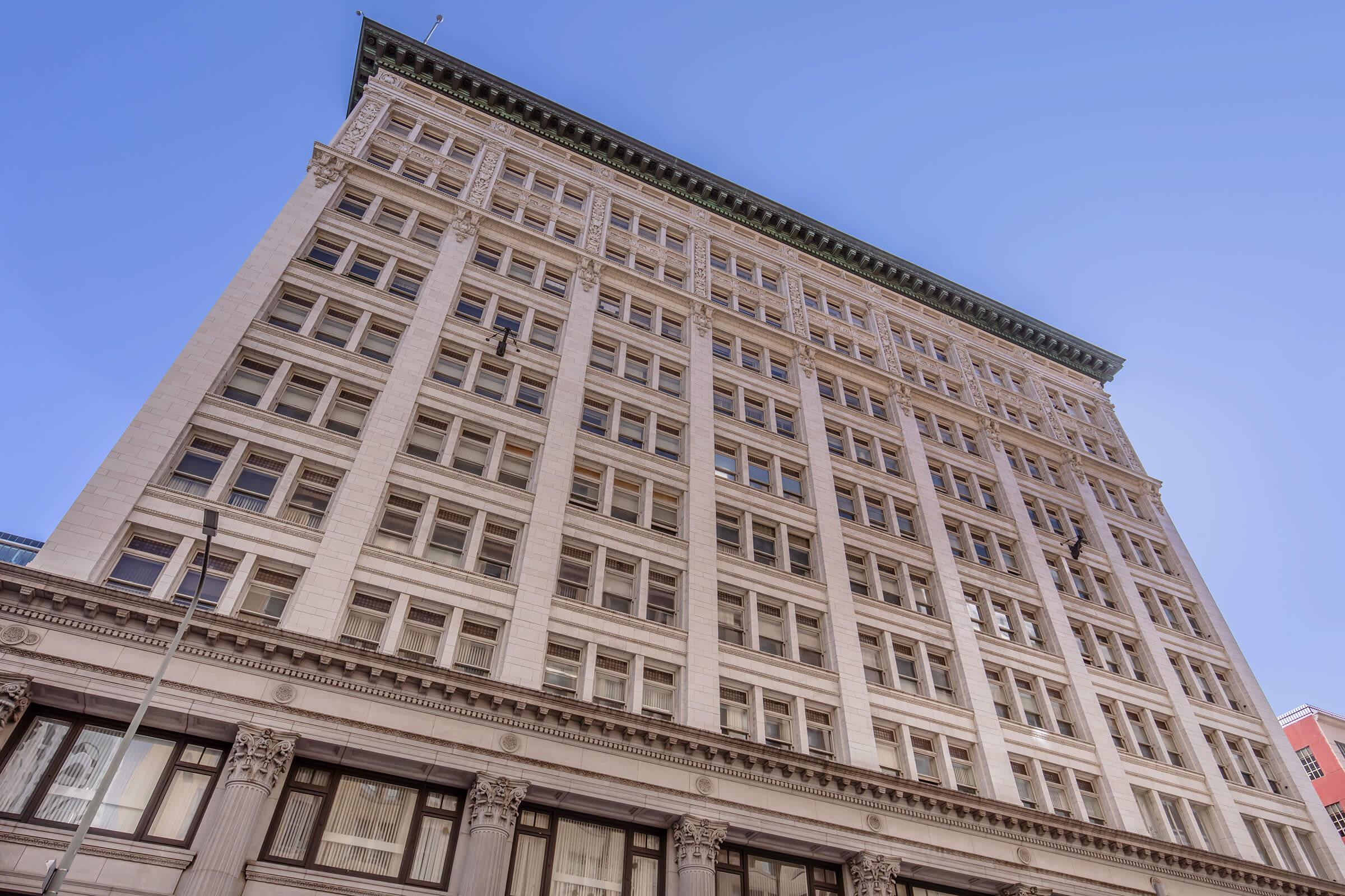 A tall historic building viewed from the ground, showcasing detailed architectural elements such as large windows and ornate columns on the lower level, against a clear blue sky. The structure features a classic style with a prominent cornice at the top.