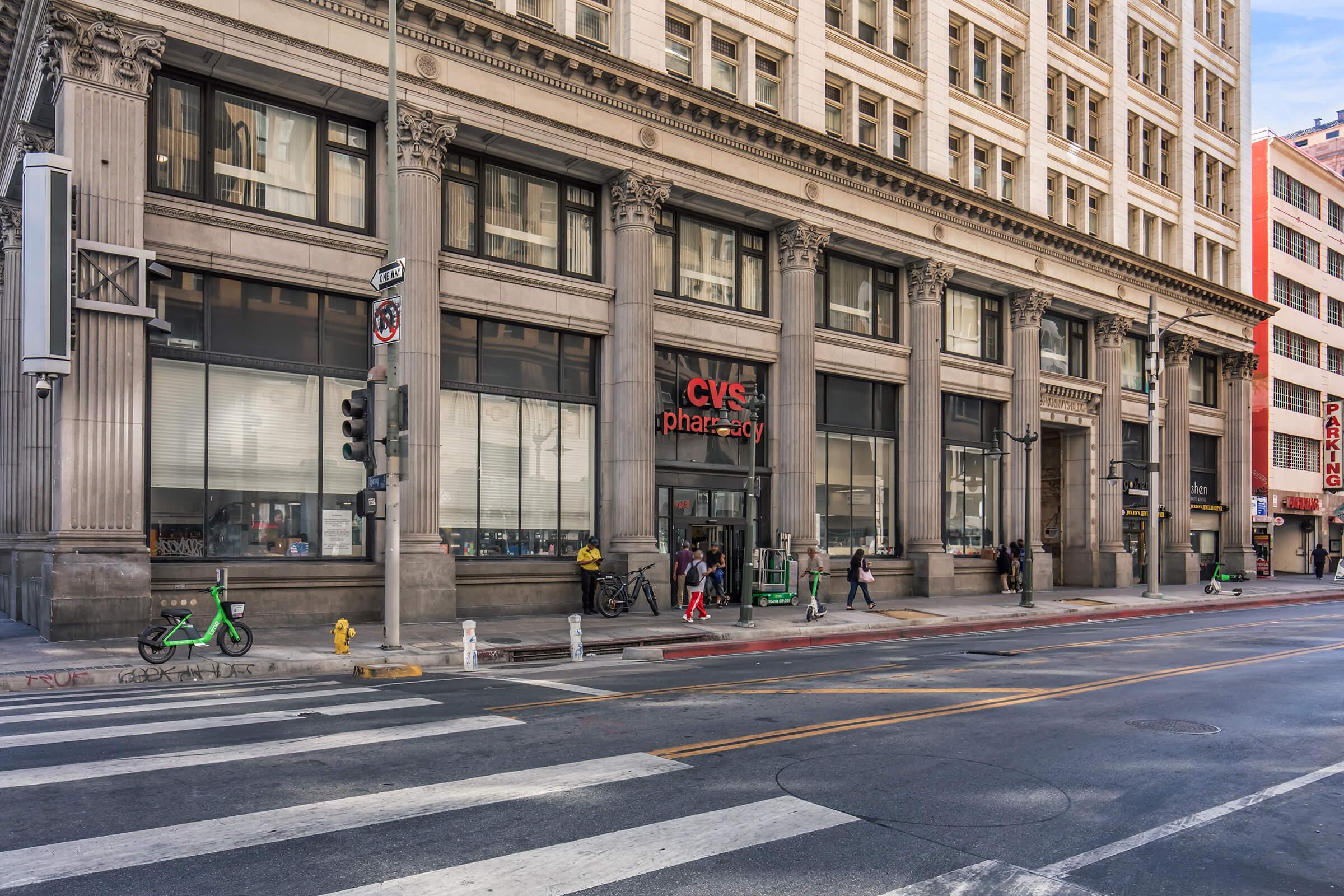 A CVS Pharmacy storefront in a city setting, featuring stone architecture and large windows. People are seen walking outside, some on bicycles, with traffic signals and crosswalks in the foreground. The scene captures the urban environment and daily life around the pharmacy.