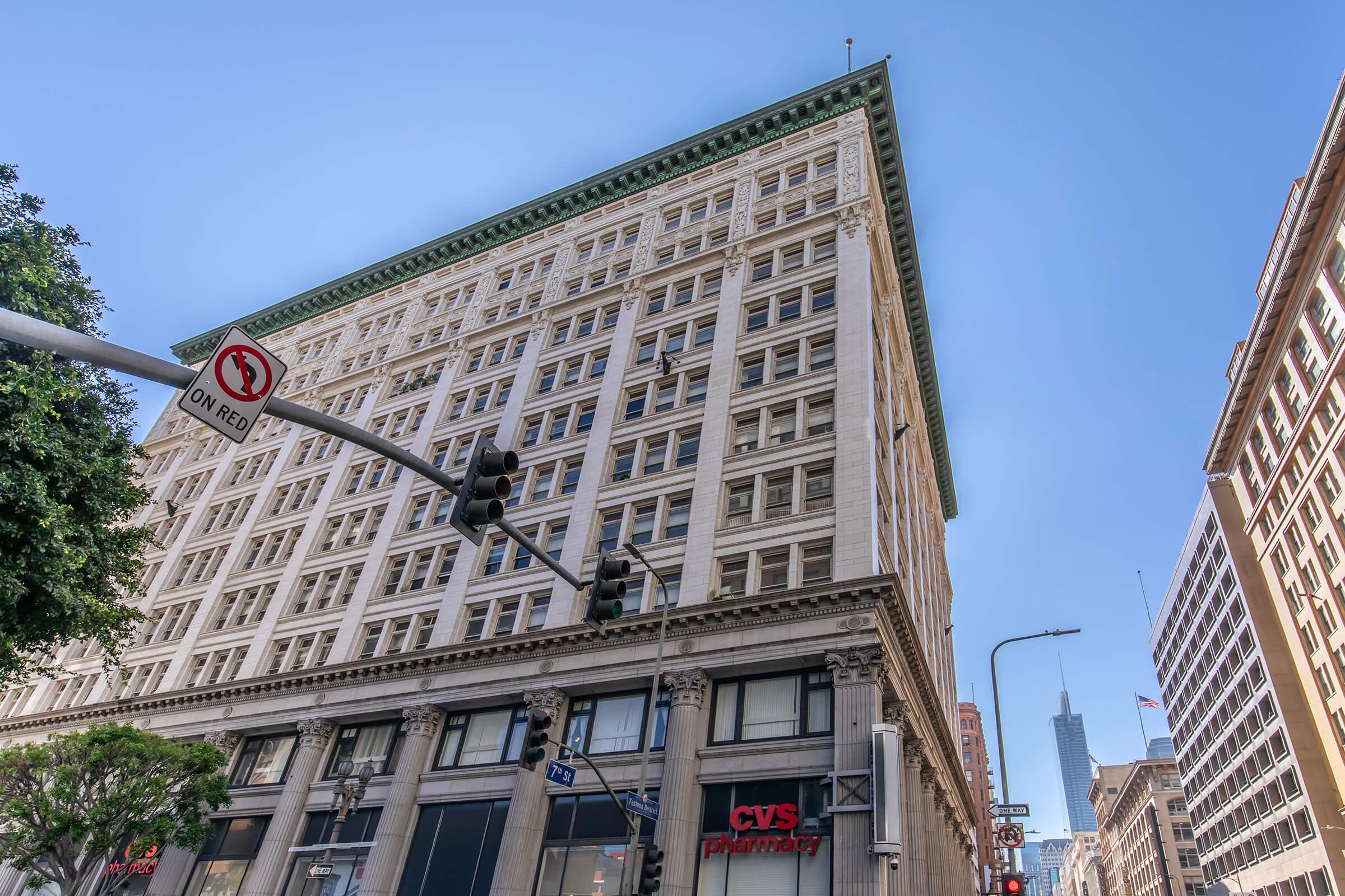 A tall historic building viewed from street level, featuring a green-topped roof and windows. A CVS Pharmacy is located on the ground floor, and a traffic signal shows "On Red." The skyline in the background includes a high-rise building, indicating an urban setting. Clear blue sky above.