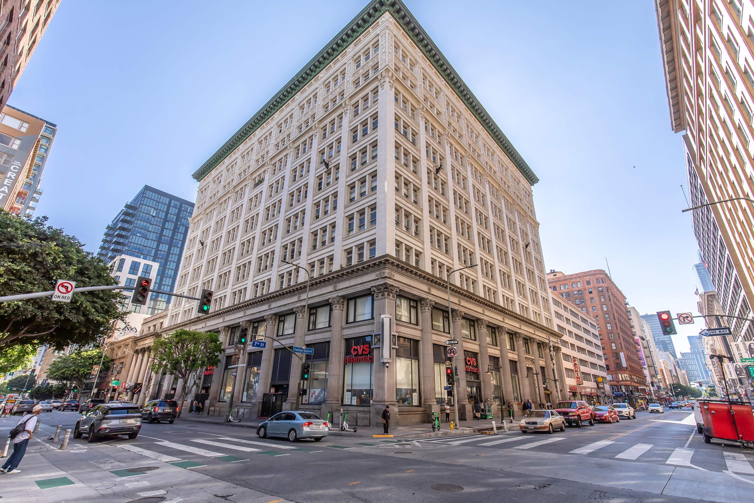 A tall, historical building with a white facade and green roof stands at a city intersection. The street features cars and pedestrians. Nearby businesses, including a CVS, are visible. Skyscrapers rise in the background under a clear blue sky.