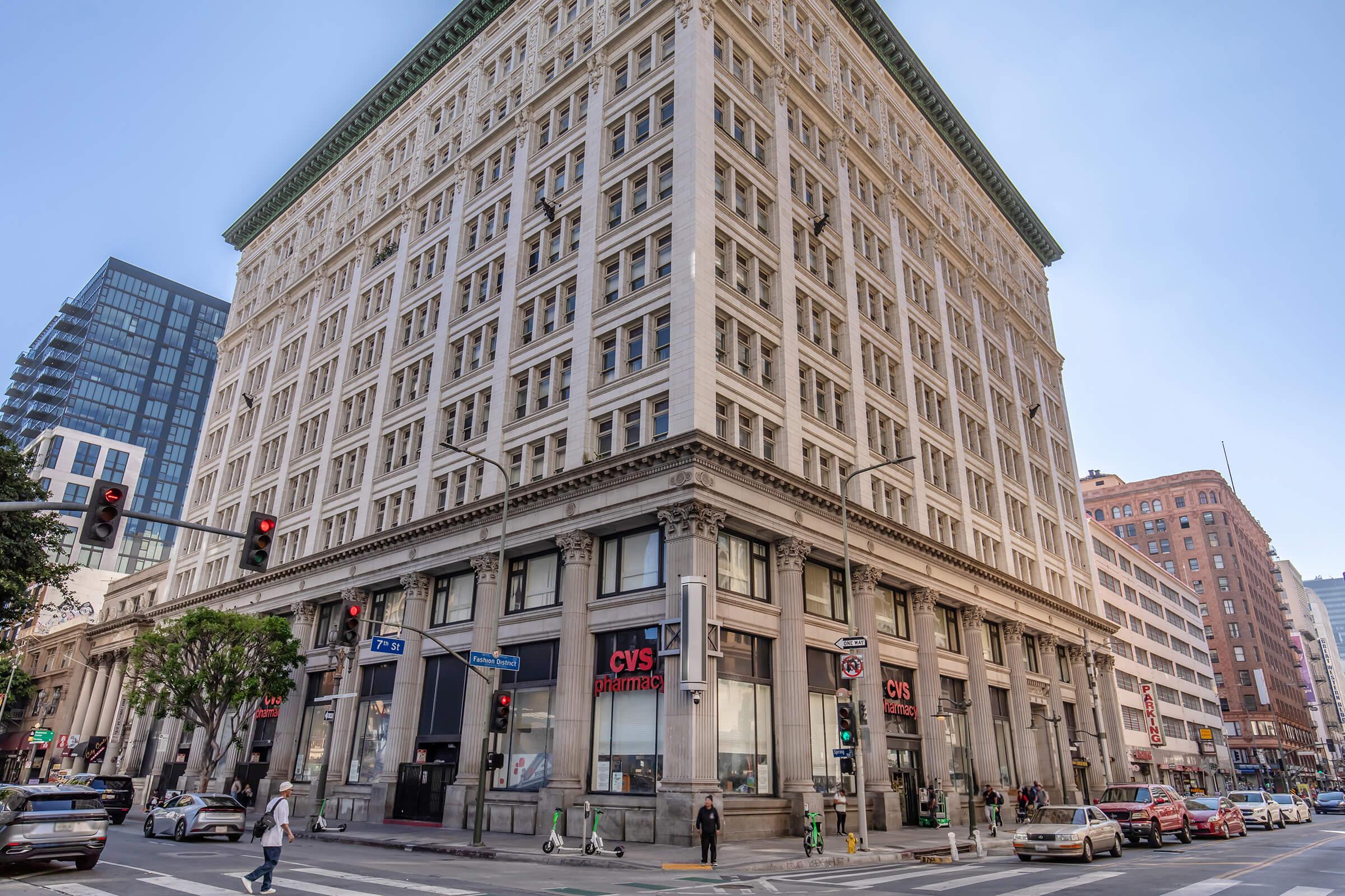 A historic building stands at the corner of an urban intersection, featuring a detailed facade with large windows. The ground floor has a CVS Pharmacy storefront. Traffic signals and pedestrians are visible, along with modern buildings in the background, indicating a bustling city atmosphere.