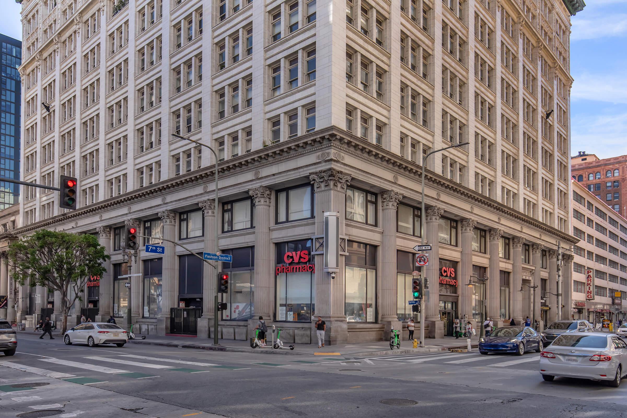 A tall, historic building with a CVS pharmacy storefront at the corner of two streets. Traffic signals are visible, along with parked and moving vehicles on the road. The scene captures an urban environment with a mix of modern and classic architecture.