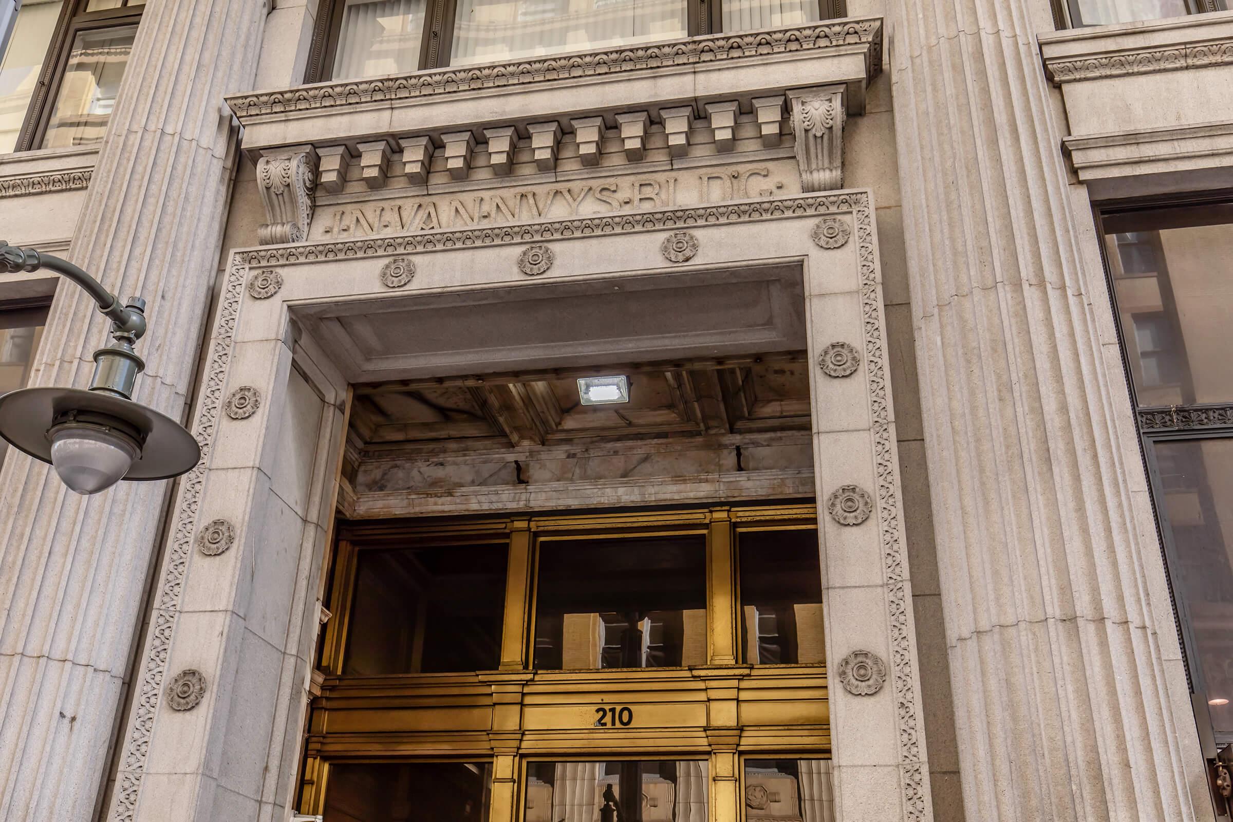 The image shows the entrance of a historic building featuring ornate columns and decorative carving. The entrance includes large, bronze doors with the number "210" visible. Above the door, architectural details and lettering are prominently displayed, contributing to the elegant design of the facade.