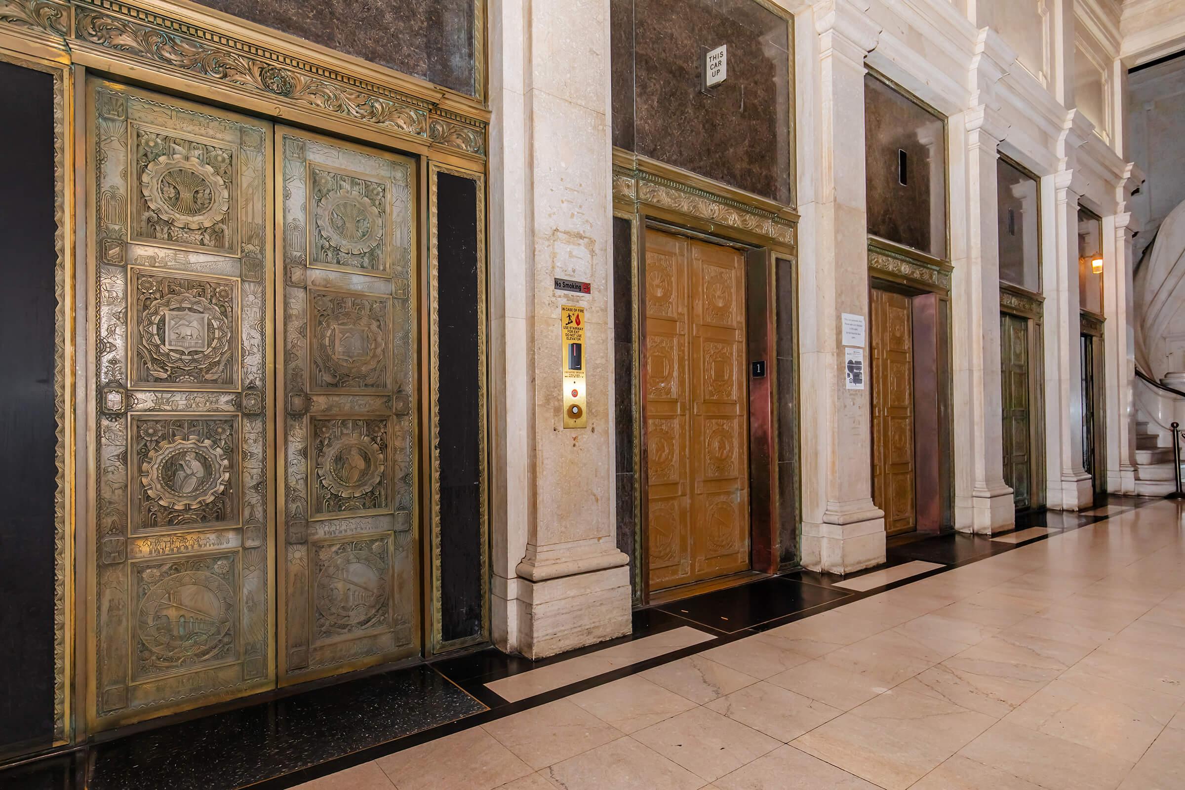 A view of ornate brass elevator doors in a historic building. The elevators feature intricate designs and detailing, with a stone wall backdrop and marble flooring. Elevators are not in use, and a call button panel is visible beside them. The overall atmosphere suggests elegance and vintage architecture.