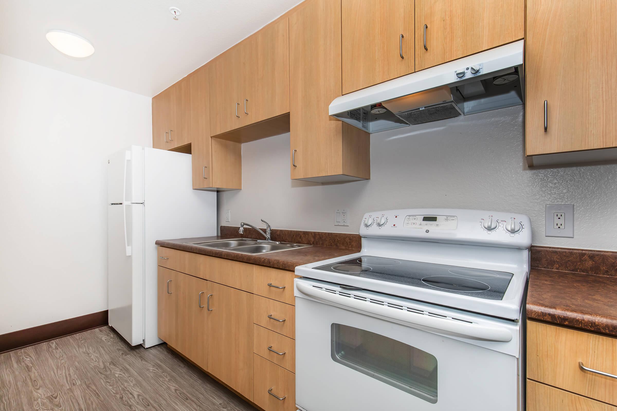 A modern kitchen featuring light wood cabinetry, a white stove with an oven, a microwave above it, and a double sink. A white refrigerator stands against the wall, and the countertops are dark brown. The floor is a wood-like laminate, and the room is well-lit with a ceiling light.