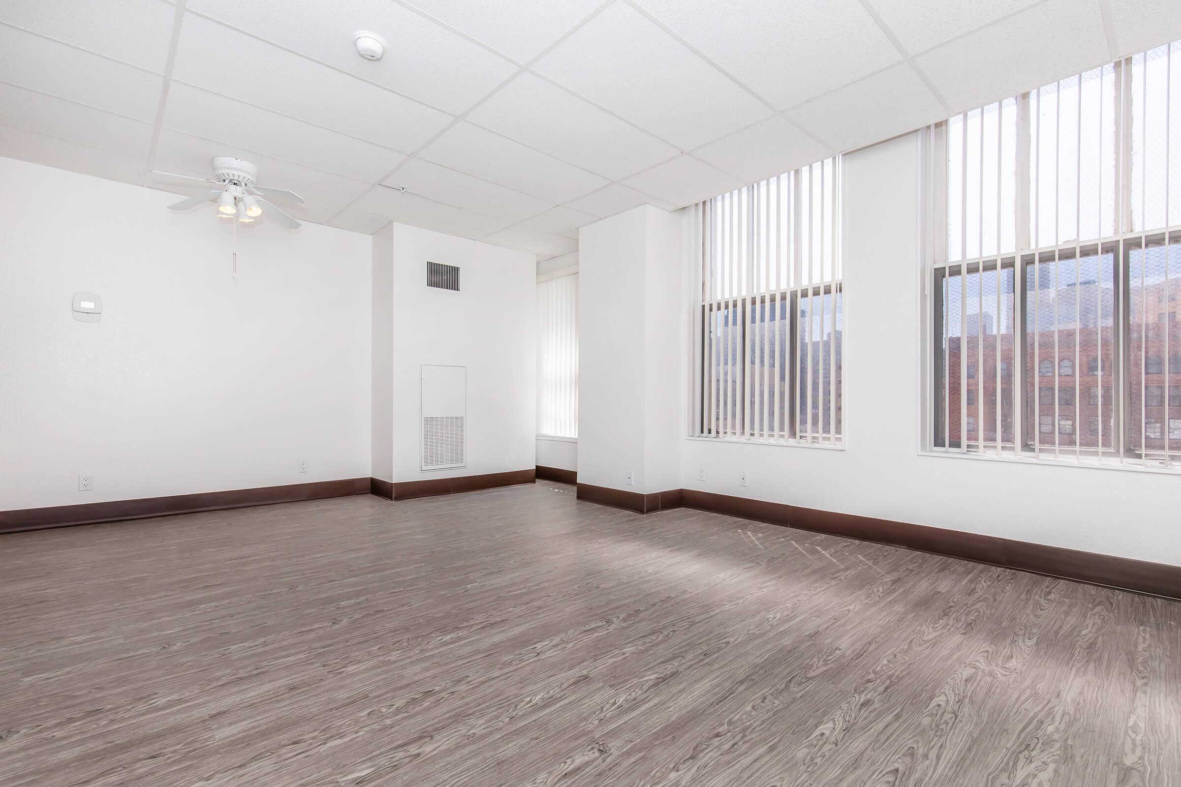 A spacious, empty room with light-colored walls and large windows featuring vertical blinds. The floor is a light, wood-like laminate. A ceiling fan is visible. The room has a clean, modern feel with ample natural light, making it suitable for various uses.