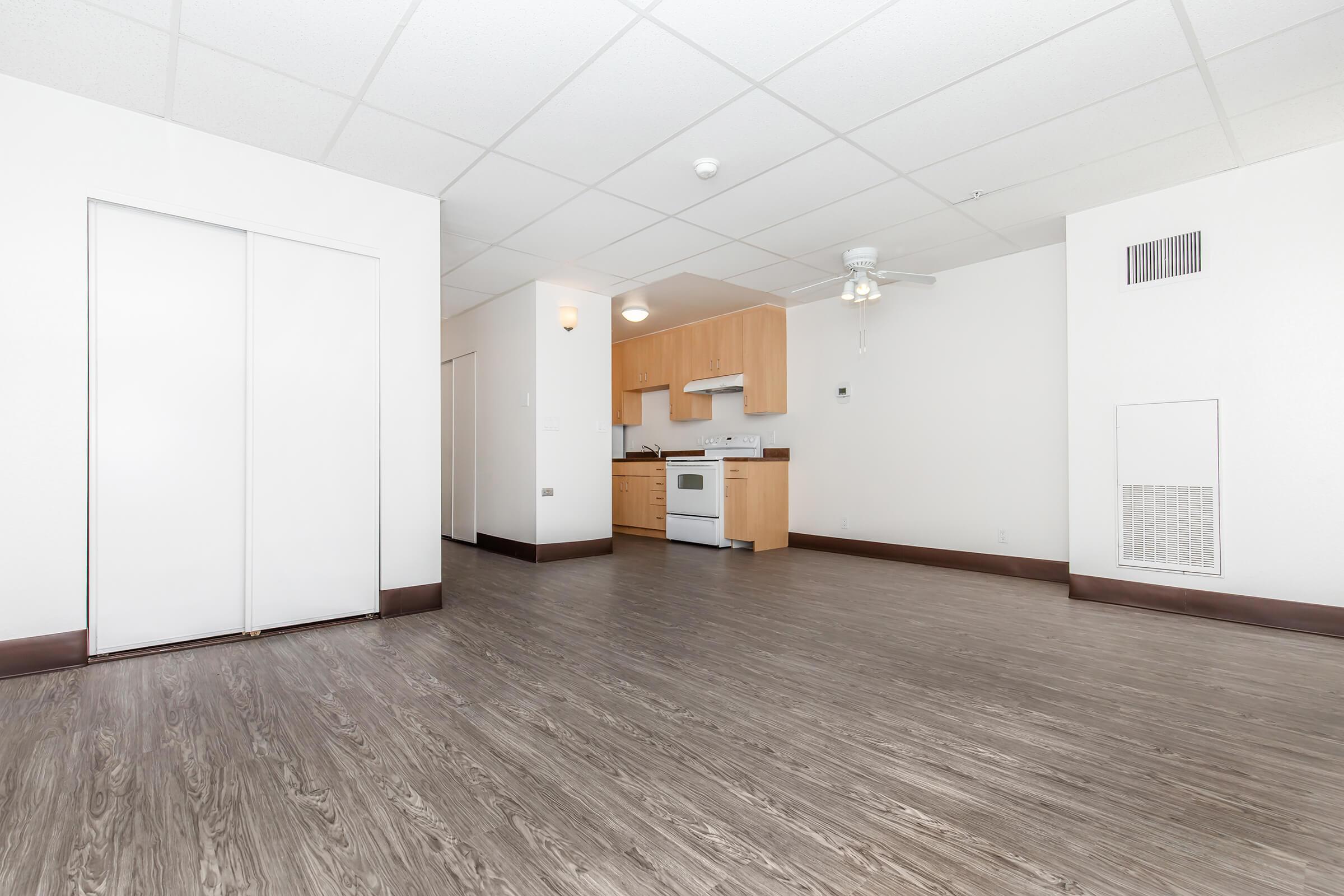 A spacious, modern studio apartment featuring an open layout with hardwood flooring. The kitchenette includes wood cabinets, a stove, and a refrigerator. There is a ceiling fan and ample natural light from large windows. The walls are painted white, and there is a closet on the left side of the image.