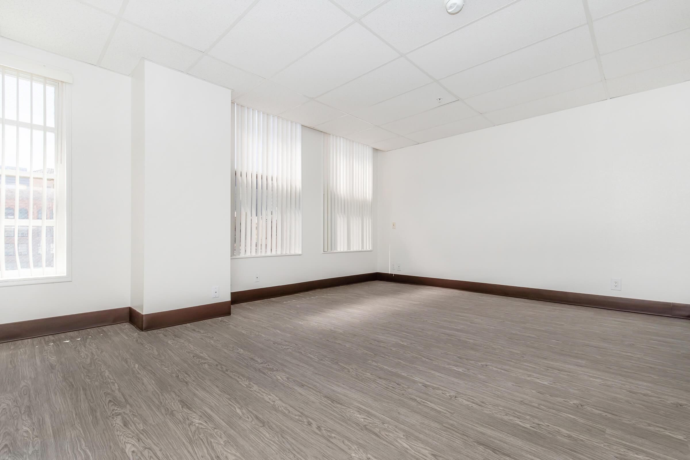 Empty interior space with light-colored walls and a laminate floor. Large windows with vertical blinds allow natural light to flood the room. The ceiling is white with a grid pattern, and there are no furnishings or decorations, creating a bright and open atmosphere.