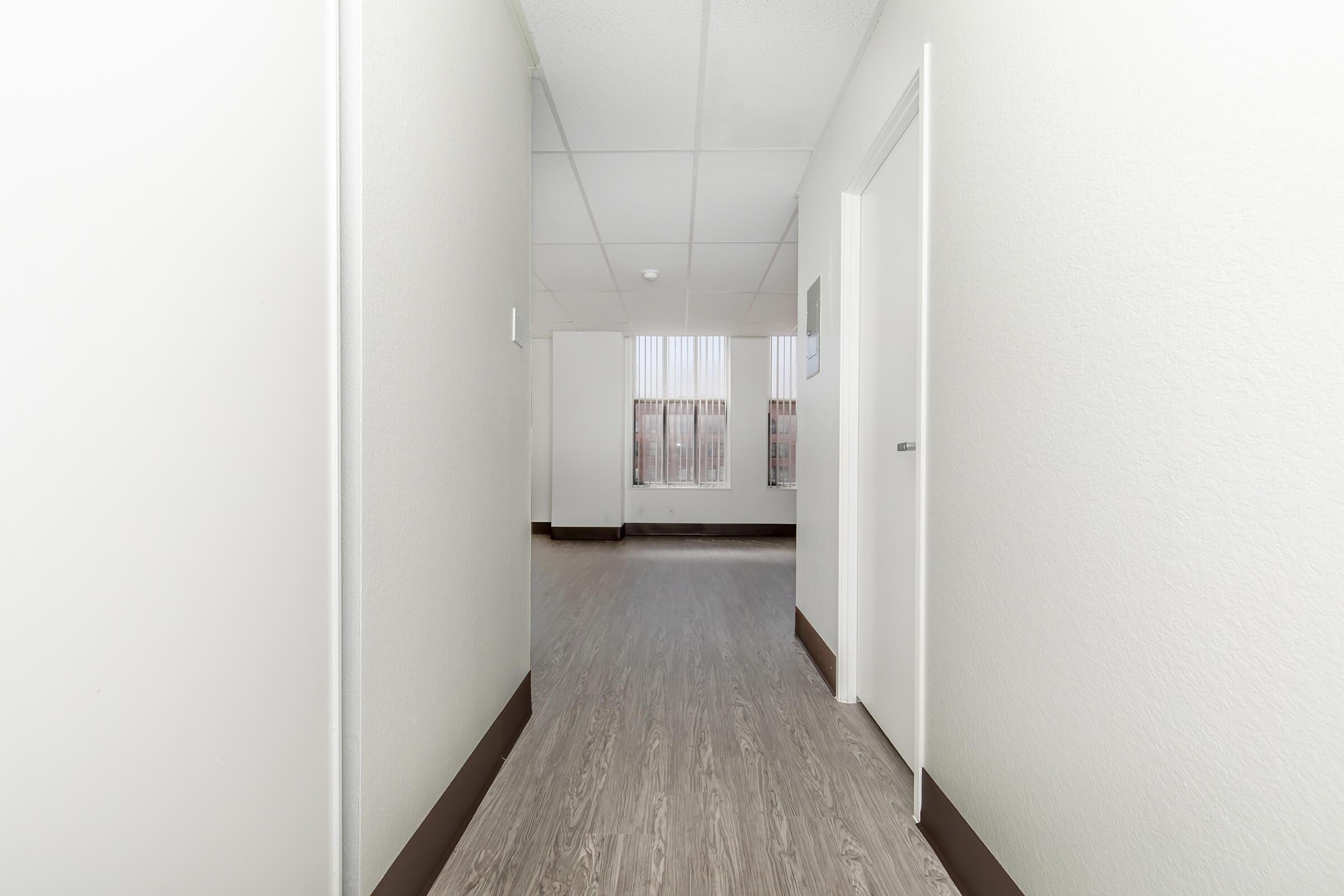 A long, narrow hallway with light-colored walls and laminate flooring. At the end of the hallway, there's a window with vertical blinds filtering natural light. White doors are visible on either side, suggesting access to different rooms or areas. The space appears clean and well-maintained.
