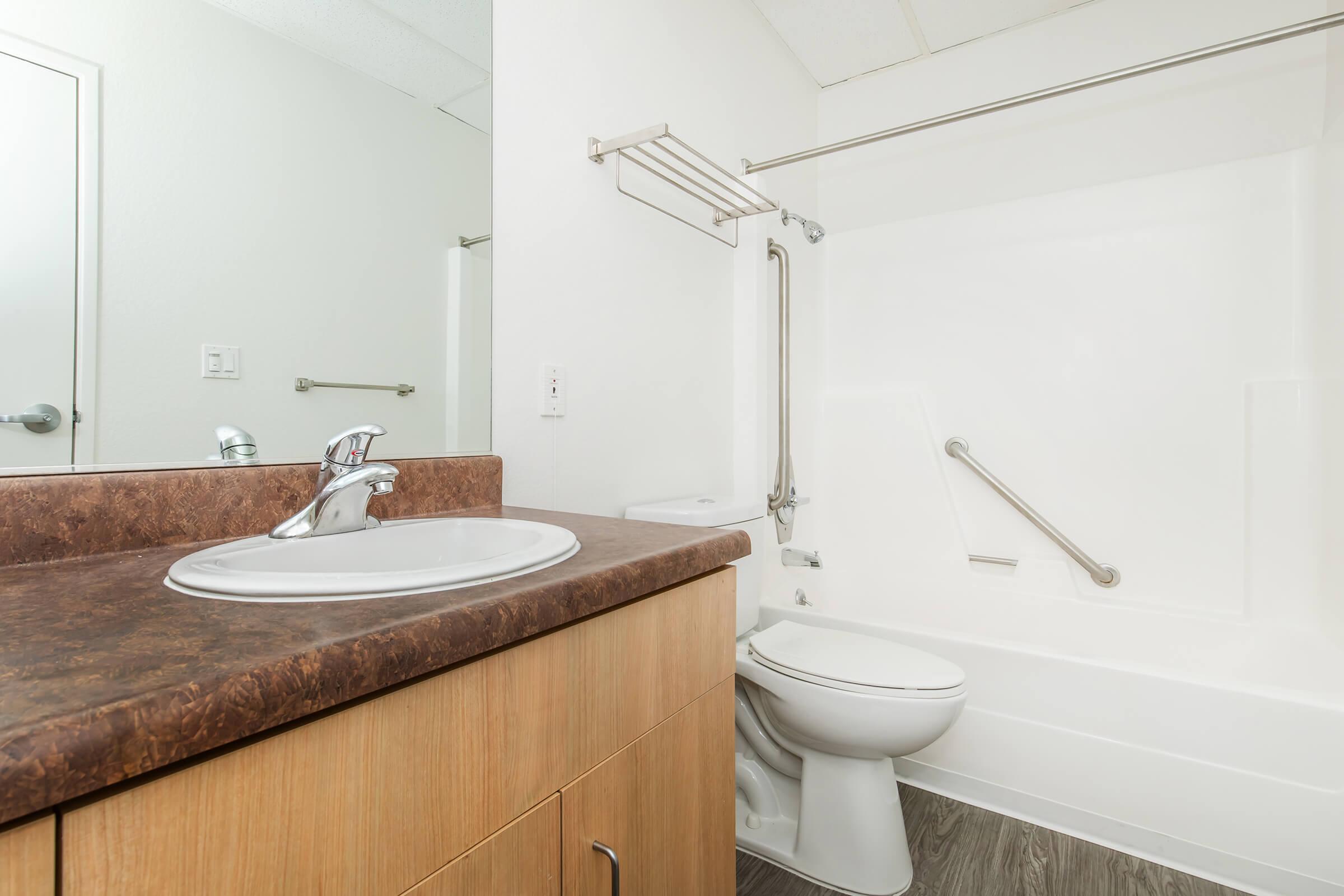 A clean bathroom featuring a modern sink with a chrome faucet, a toilet, and a white bathtub with grab bars. The walls are painted white, and there's a wooden cabinet under the sink. The flooring appears to be vinyl, giving the space a bright and spacious feel.