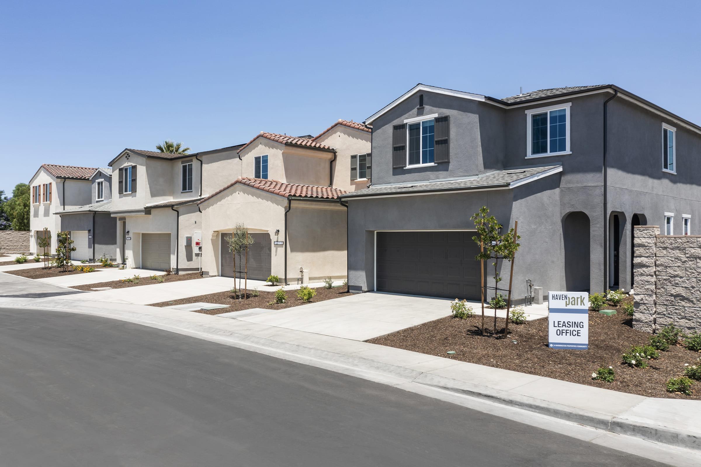 A row of modern residential houses with stucco exteriors and varied architectural styles. The street is lined with new homes, some featuring attached garages. A sign indicates the presence of a leasing office. The scene is set under a clear blue sky, highlighting the suburban neighborhood's appeal.