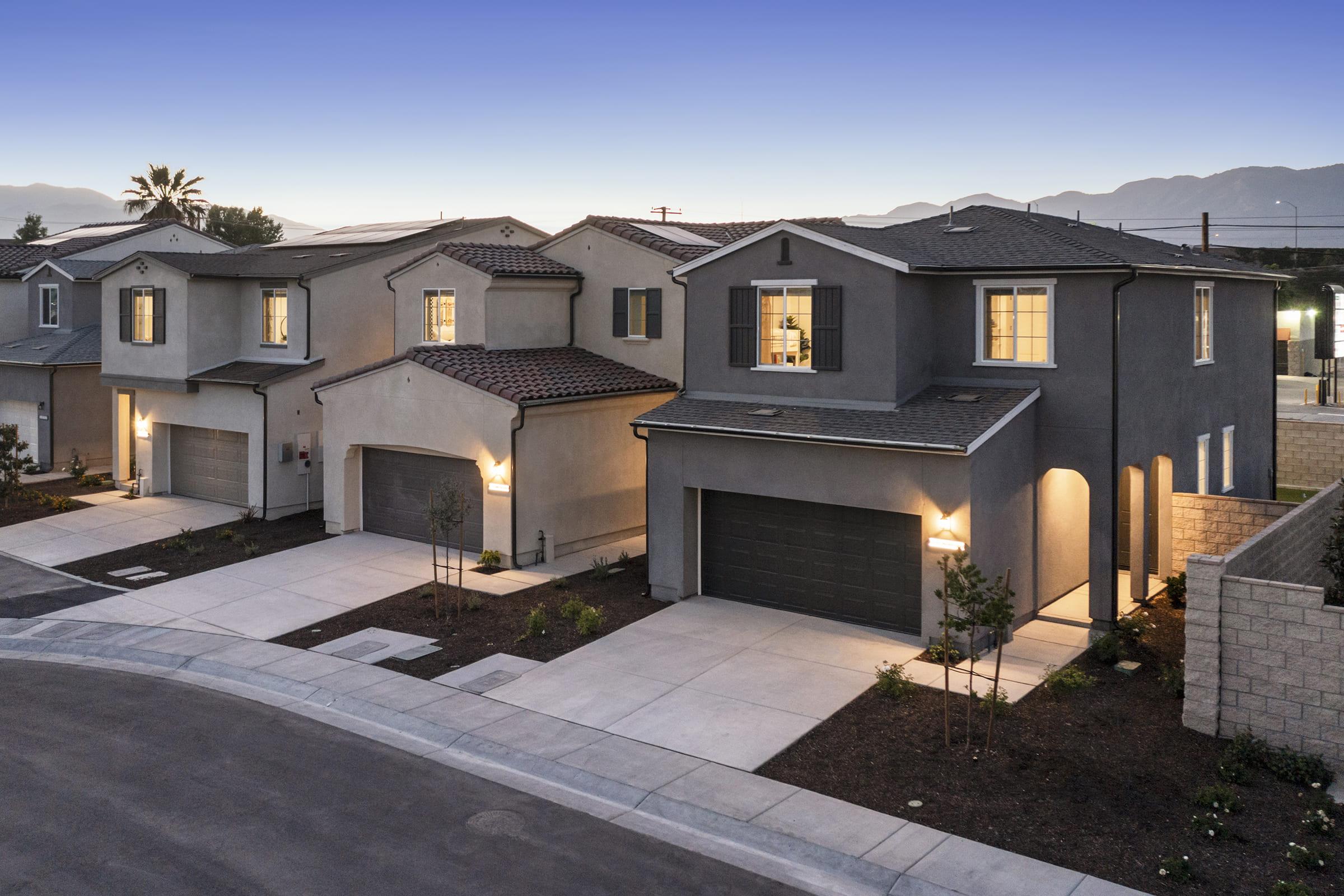A modern residential neighborhood at dusk, featuring two stylish houses with gray exteriors and tiled roofs. The homes have well-lit garages and soft exterior lighting, with neatly landscaped walkways and a quiet suburban street lined with young trees in the foreground. Mountains are visible in the background under a clear blue sky.