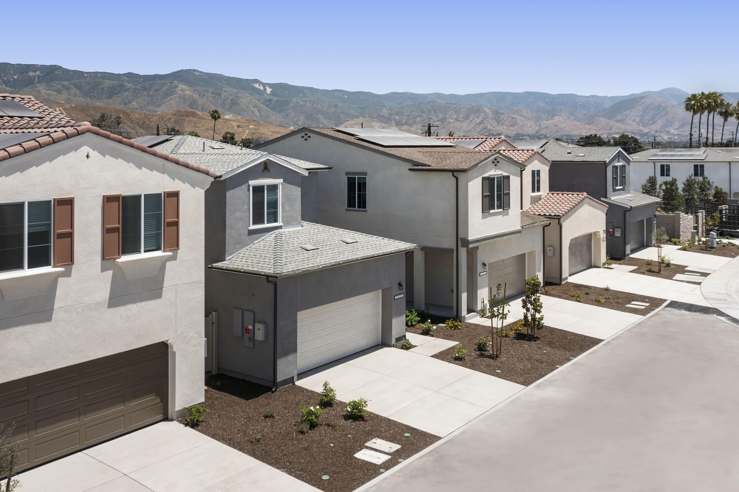 A row of modern residential houses with a mix of architectural styles, featuring garages, driveways, and landscaped yards. In the background, rolling hills and a clear blue sky enhance the suburban setting. The scene conveys a peaceful, well-maintained neighborhood.