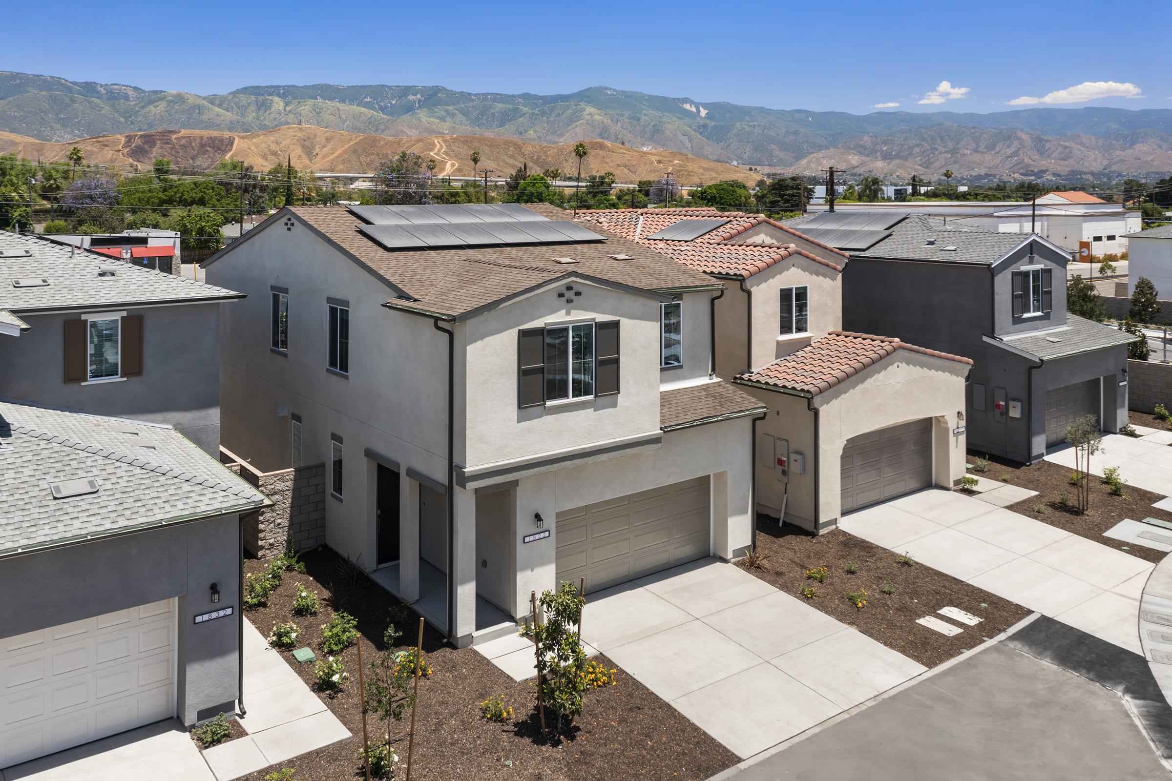 Aerial view of modern two-story houses in a suburban area, featuring various architectural styles. The foreground shows a new development with landscaped yards and driveways. In the background, mountains are visible under a clear blue sky, indicating a sunny day. Solar panels are installed on some roofs.