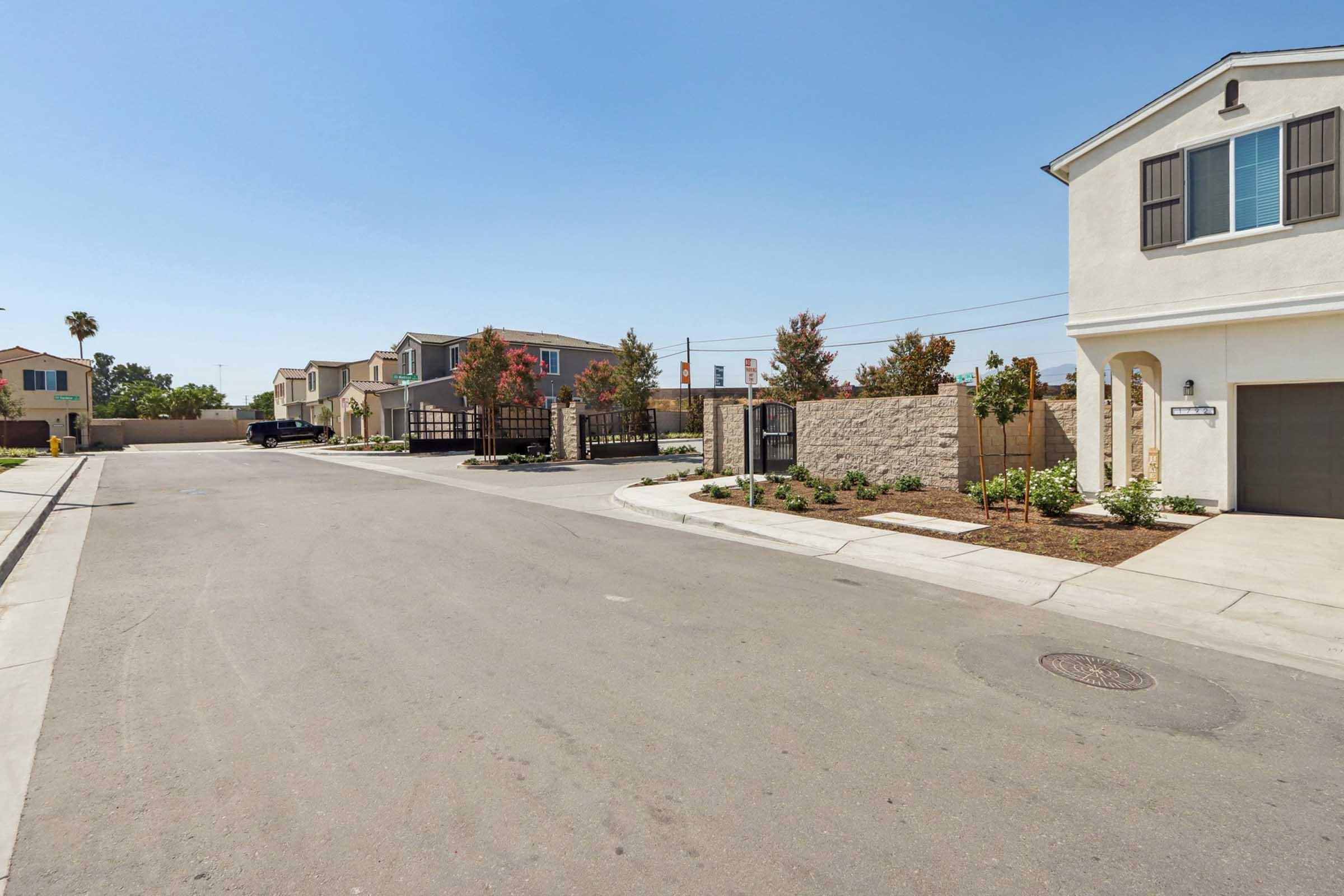 A quiet residential street with single-family homes on the right and a gated entrance on the left. The road has green landscaping on the sides and is bathed in bright sunlight under a clear blue sky. There are no visible pedestrians or vehicles on the street.