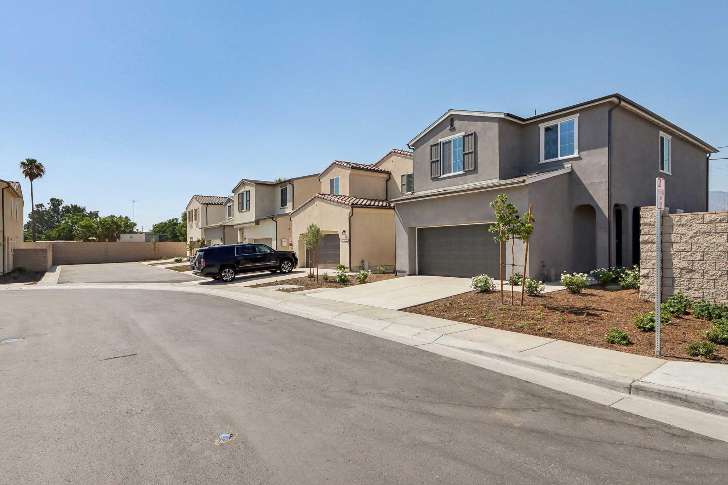 A quiet residential street with several two-story houses, featuring a driveway with a parked black SUV. One house has a landscaped front yard with small plants, while another house is partially visible in the background. Clear blue sky and a palm tree can be seen nearby.