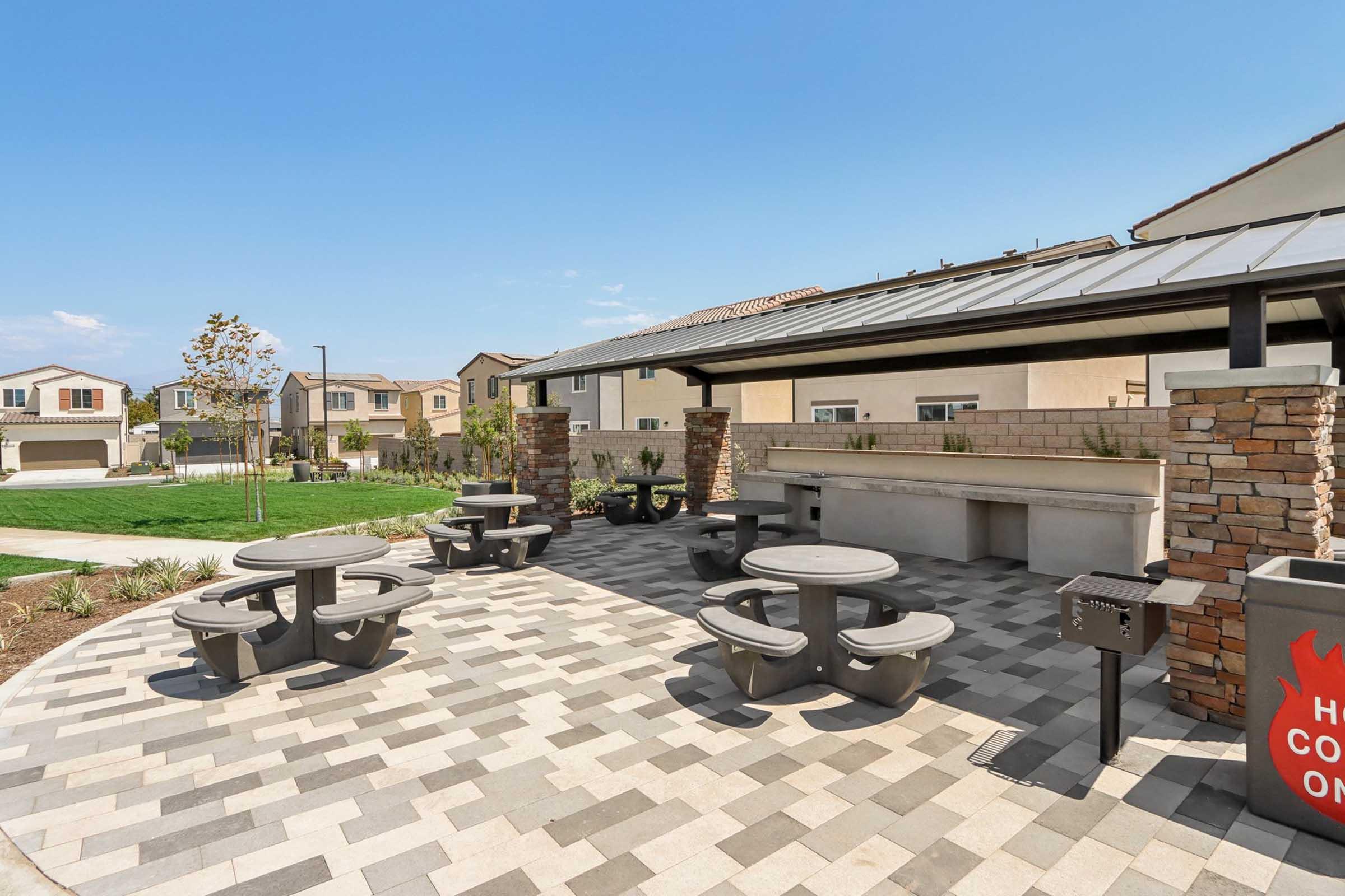 An outdoor community area featuring gray picnic tables and chairs on a patterned stone patio. There is a shaded pavilion with a bar-style counter and landscaping in the background. A grassy area and residential houses are visible in the distance under a clear blue sky.