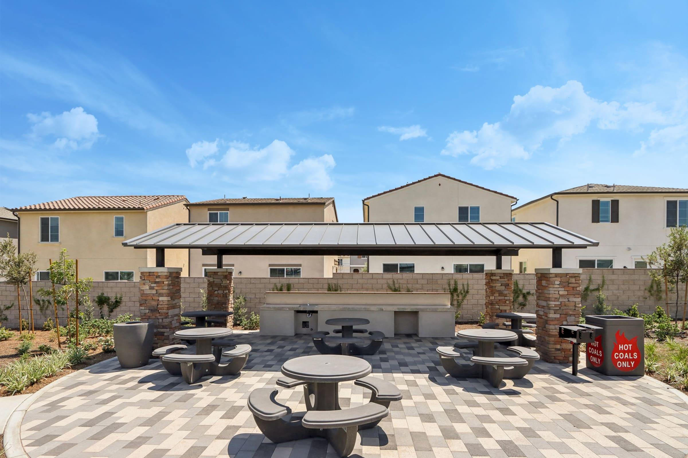 An outdoor picnic area featuring several round concrete tables and benches under a covered pavilion. The space is surrounded by neatly landscaped grounds, with a grill station and a trash bin. Residential houses are visible in the background under a clear blue sky.