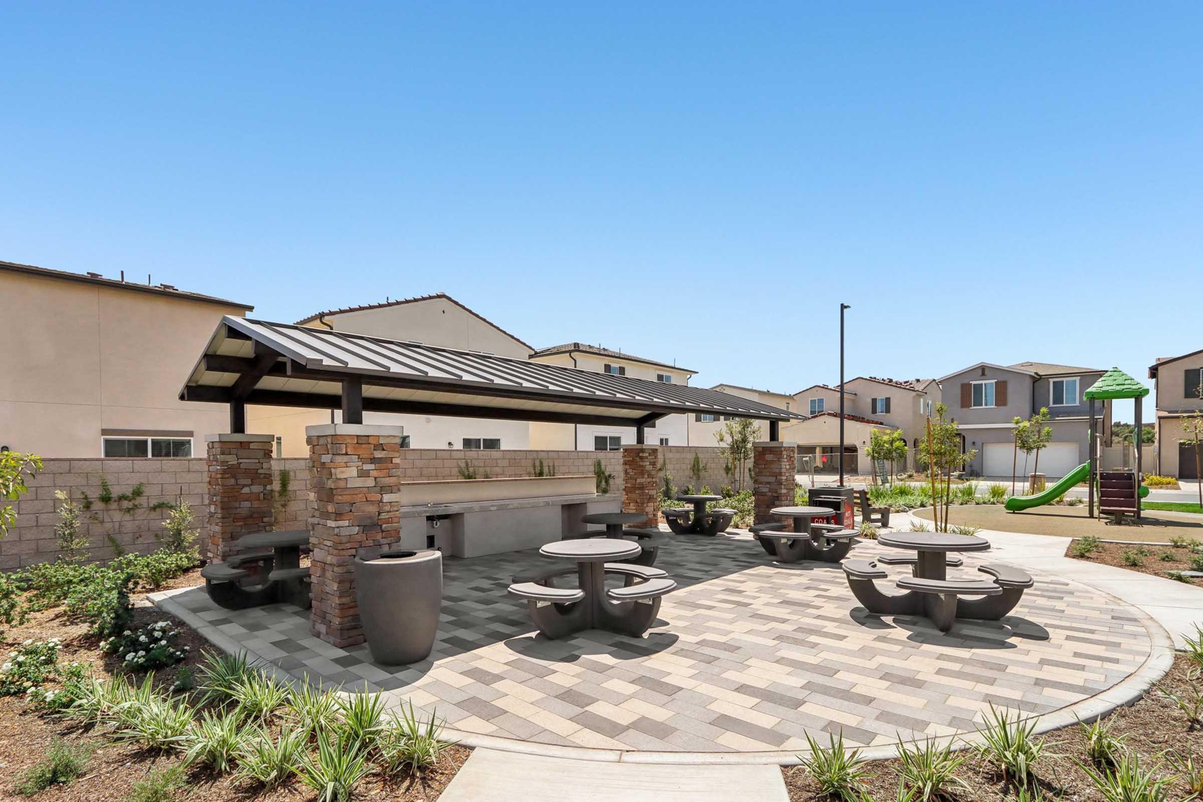 A spacious outdoor picnic area featuring stone tables and seating under a covered structure. The surrounding landscape includes paved pathways, greenery, and residential buildings in the background, creating a welcoming environment for gatherings and relaxation. Clear blue sky overhead.