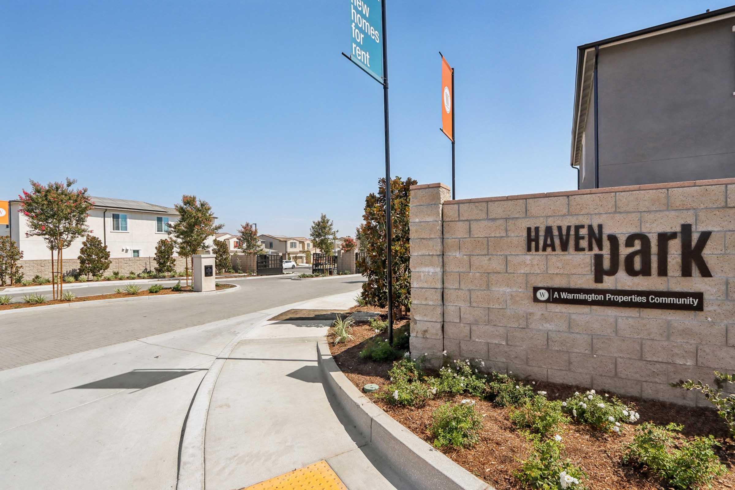 Entrance to Haven Park, a residential community, featuring a welcoming sign with modern design. The surrounding area includes paved roads and landscaped greenery, with flags indicating community information. Bright sky in the background suggests clear weather.