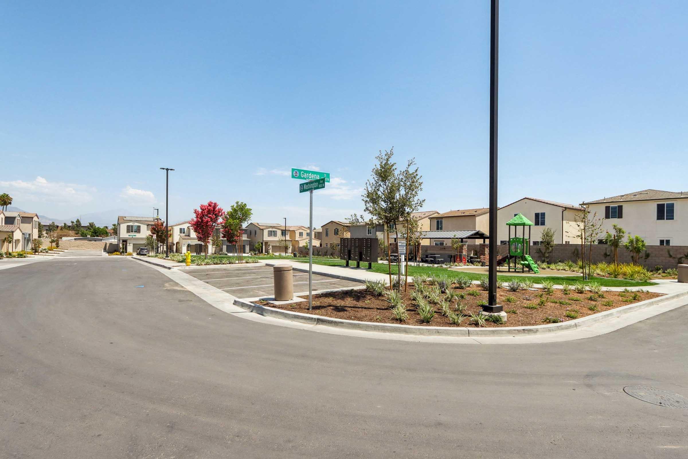 A quiet residential street with new houses lining both sides. The foreground shows a circular intersection with a street sign for "Cypress Street." There are patches of landscaped greenery, a small playground visible in the distance, and blue skies overhead.