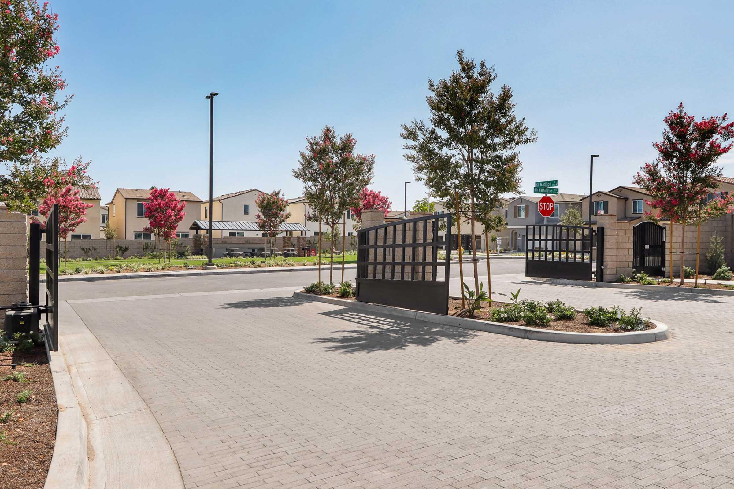 A view of a residential entrance featuring a stylish black gate, lined with trees and manicured landscaping. Bright blue skies above, and a stop sign visible in the distance. The area includes modern houses in a planned community with well-maintained pathways and greenery.