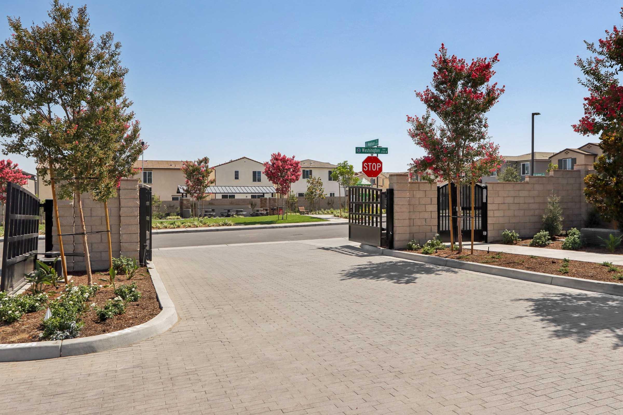 A gated entrance with a stop sign, leading into a residential area. Lush green landscaping and trees with red leaves are visible on both sides of the entrance. Modern homes can be seen in the background under a clear blue sky. The pavement is made of bricks, creating a welcoming atmosphere.