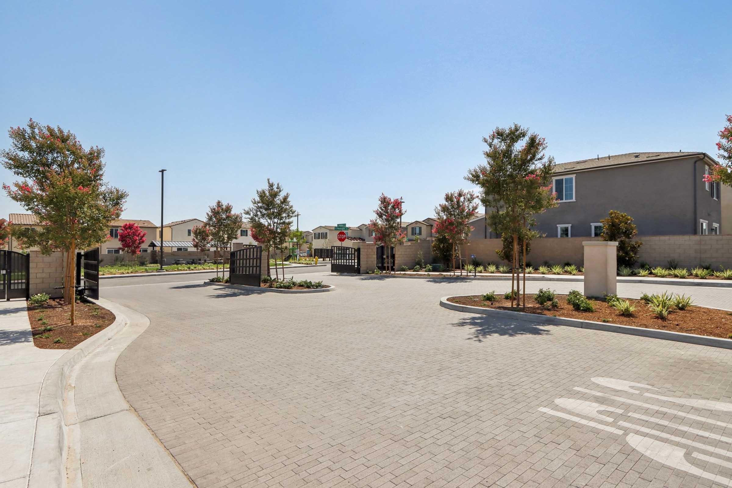 A view of a gated community entrance featuring a paved road, landscaped trees, and houses in the background. The clear blue sky adds to the bright atmosphere, while the gated entry points on either side invite residents into the neighborhood.