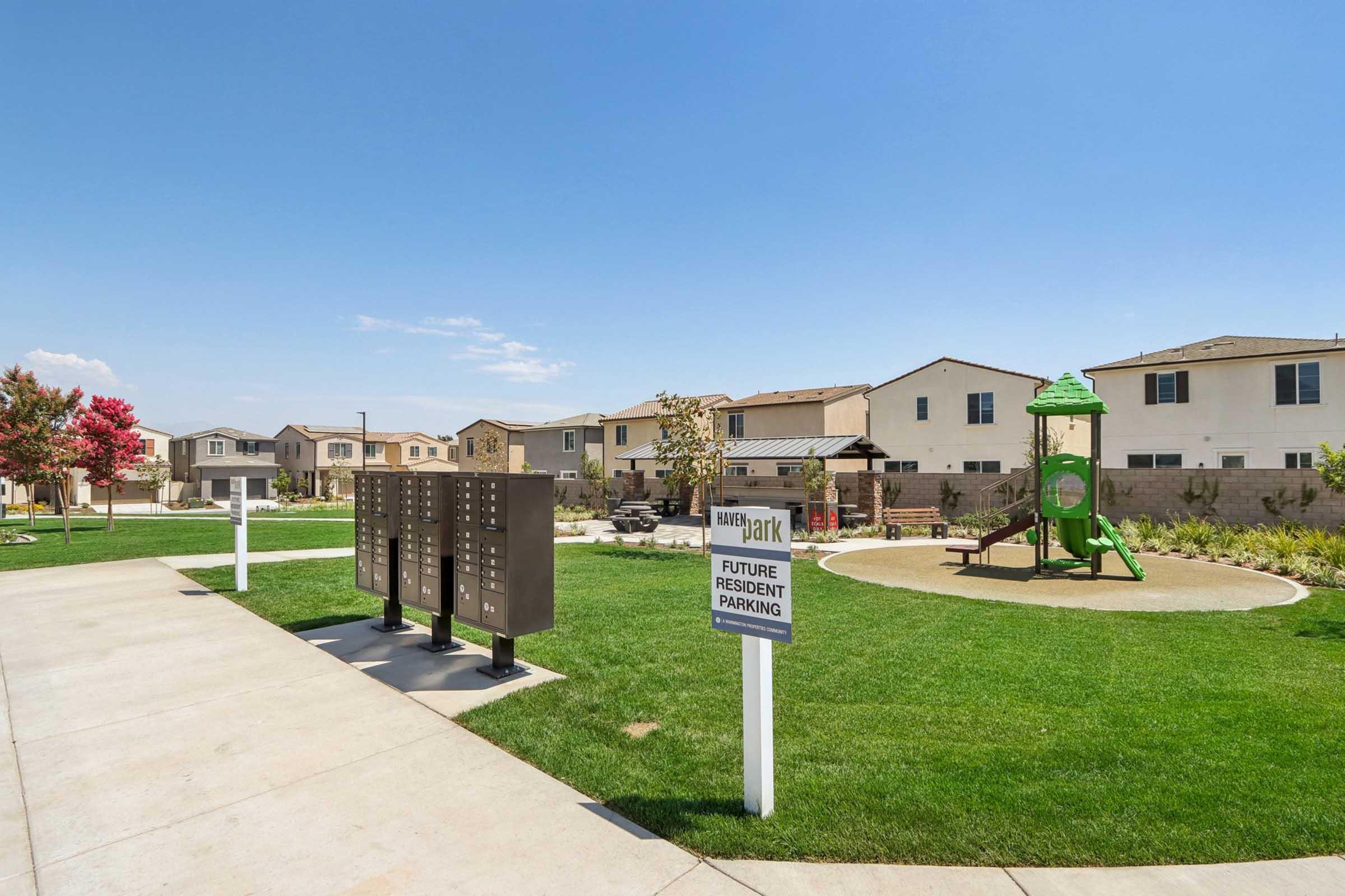 View of a residential community featuring a playground, grassy area, and mailboxes. New homes are visible in the background, with a sign indicating future resident parking. The sky is clear, and trees with autumn-colored leaves add to the landscape.