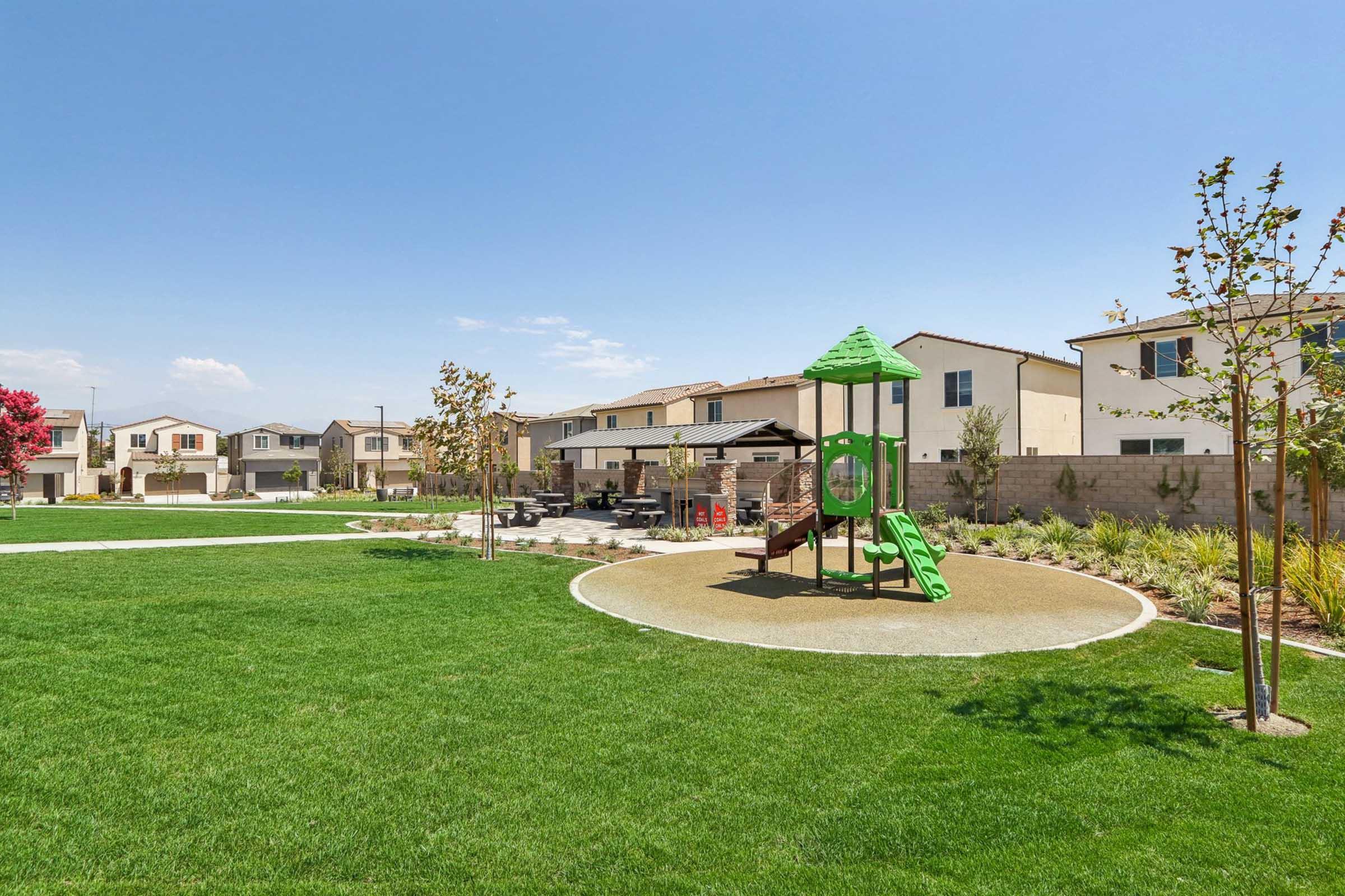 A bright green playground structure with a slide and climbing features is situated on manicured grass in a sunny park. The background features several modern residential houses, and small trees are planted around the area, creating a welcoming outdoor space for families and children.