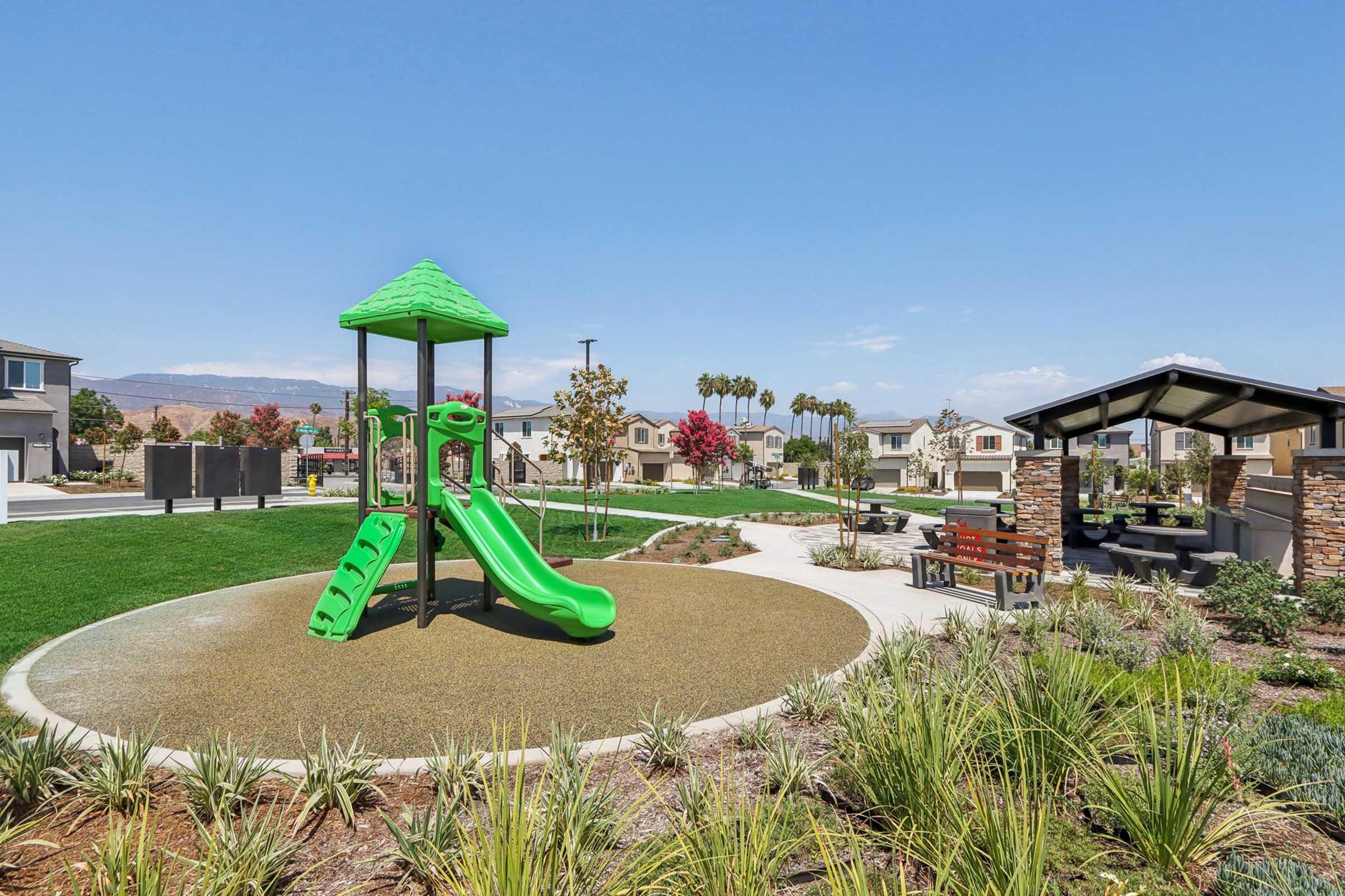 A bright green playground with a slide and climbing structure, surrounded by lush landscaping and trees. In the background, there are modern houses and a shaded seating area with stone pillars. The sky is clear and blue, creating a vibrant, family-friendly atmosphere.