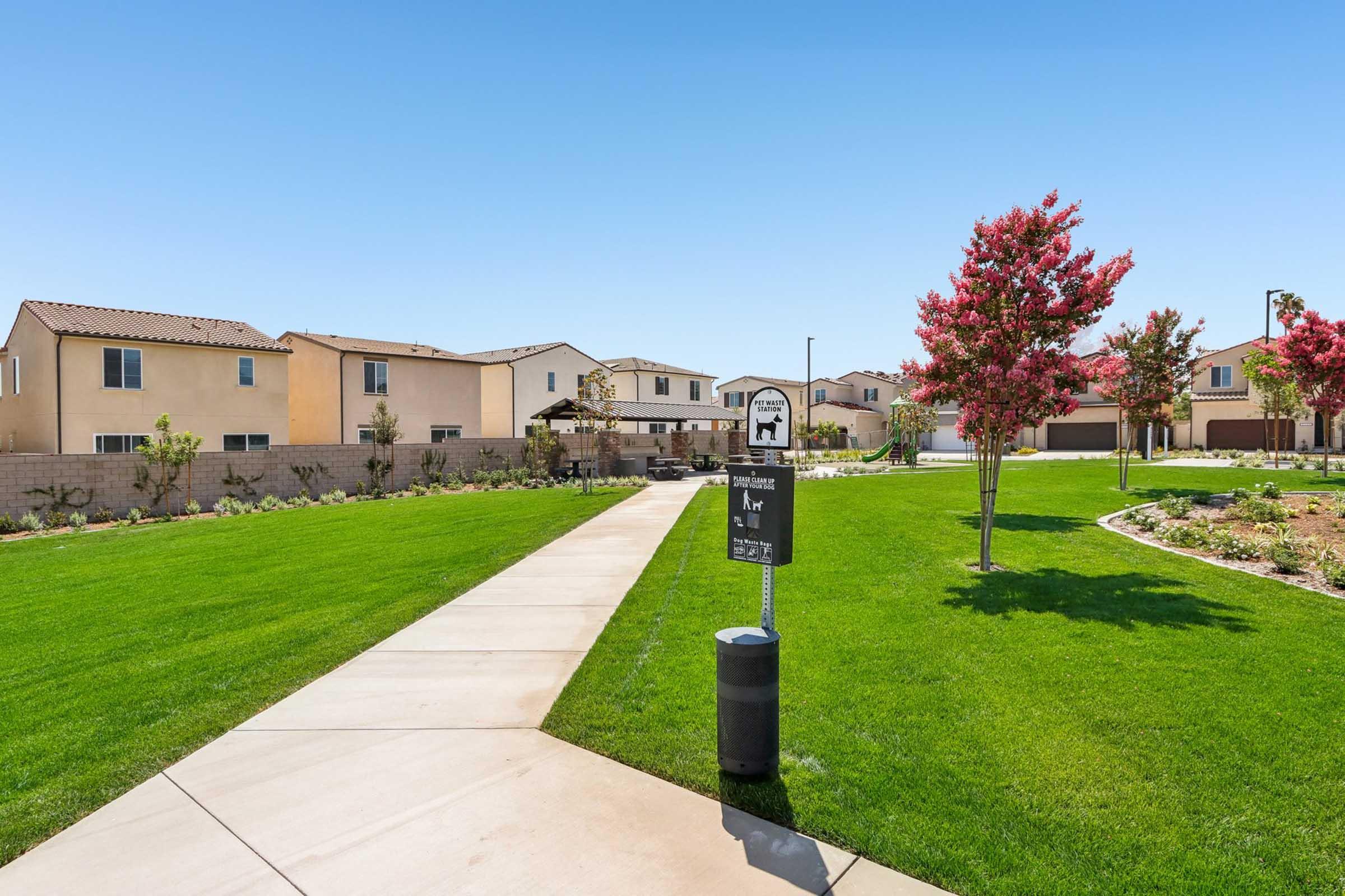 A well-maintained residential area featuring a green lawn, a sidewalk winding through the neighborhood, and newly built houses in the background. Decorative flowering trees are planted along the path, enhancing the community's charm. A sign indicating a pet park is visible along the walkway.