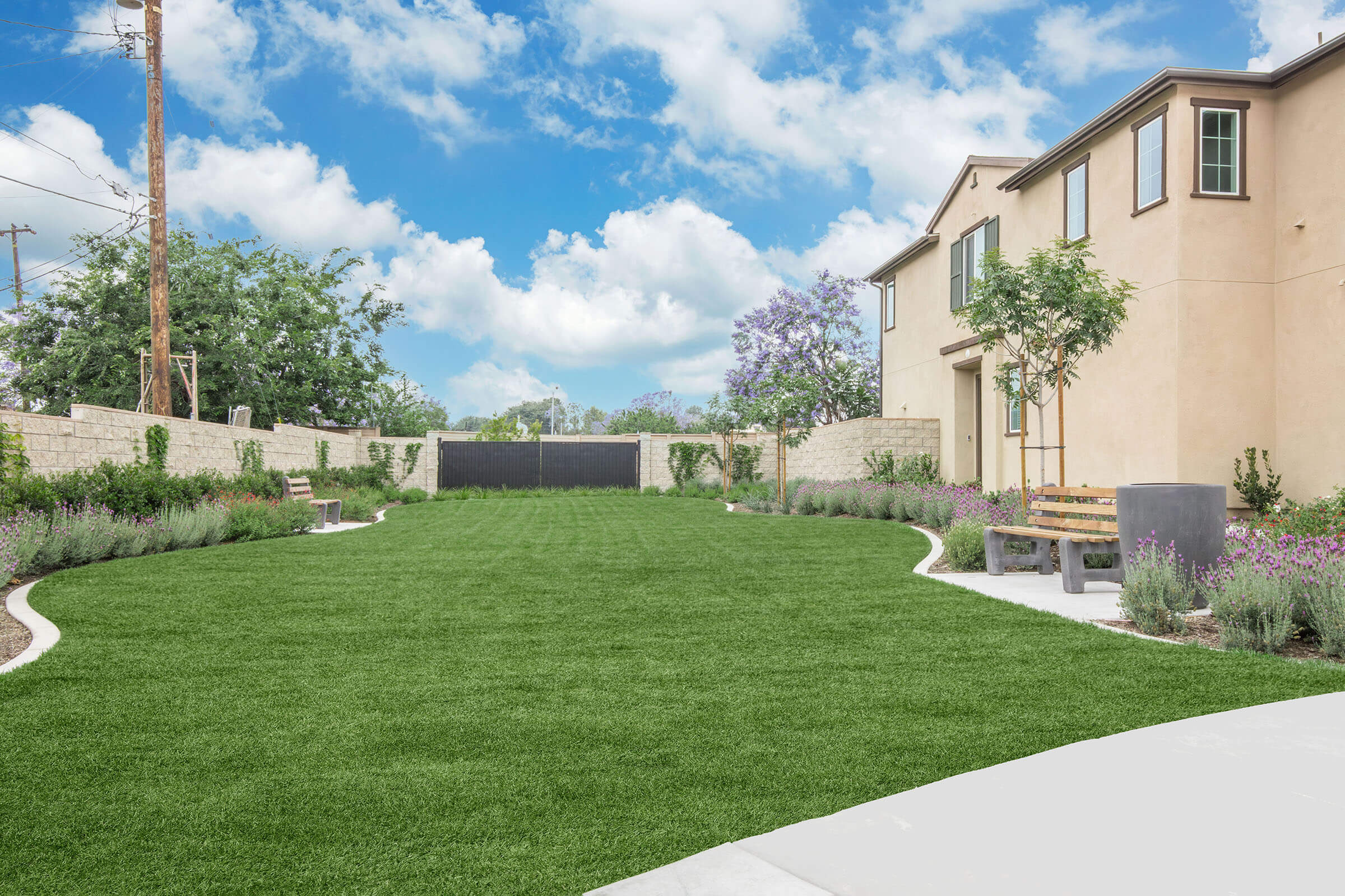 A spacious, well-maintained backyard featuring lush green grass, flowering plants along pathways, a wooden bench, and a stone wall. In the background, there are two houses, with a clear blue sky and fluffy clouds overhead, creating a serene outdoor setting.