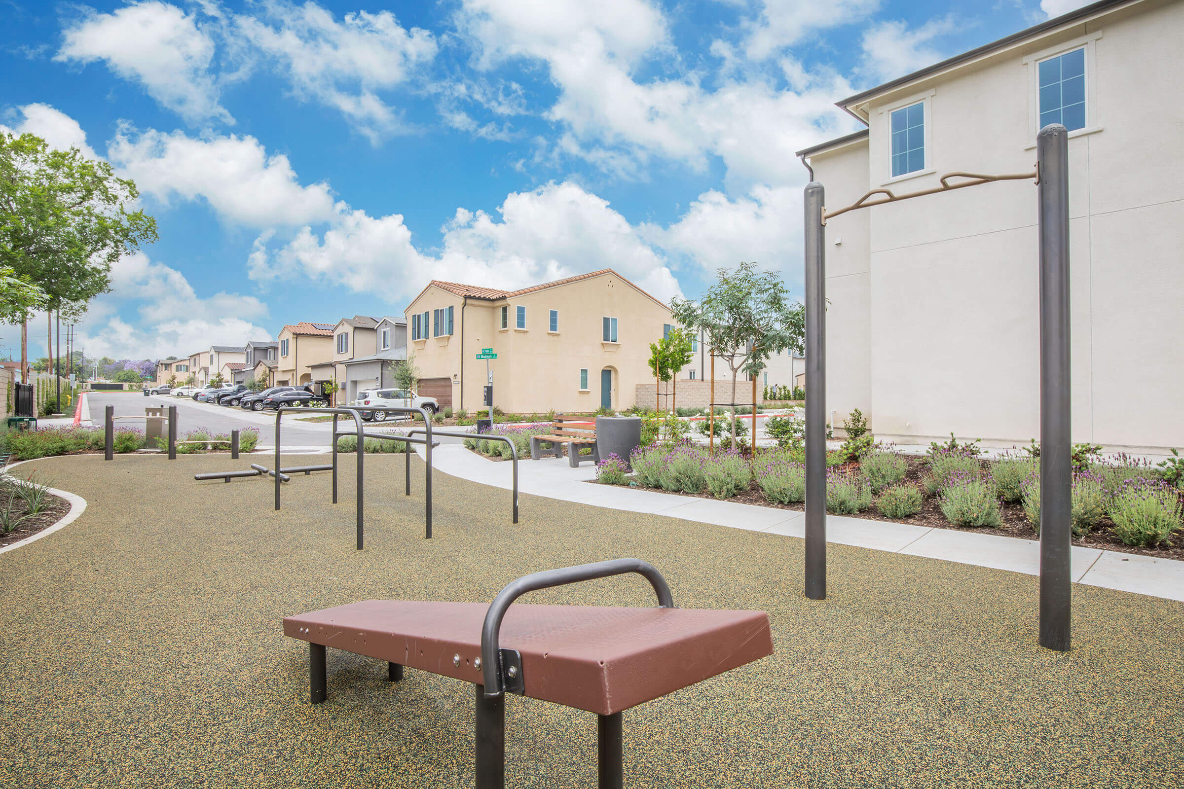 A modern outdoor fitness area with exercise equipment, including a bench and pull-up bars, surrounded by landscaped green spaces. In the background, there are residential buildings and a clear blue sky with scattered clouds. The pathway is lined with shrubs and trees, creating a welcoming atmosphere.