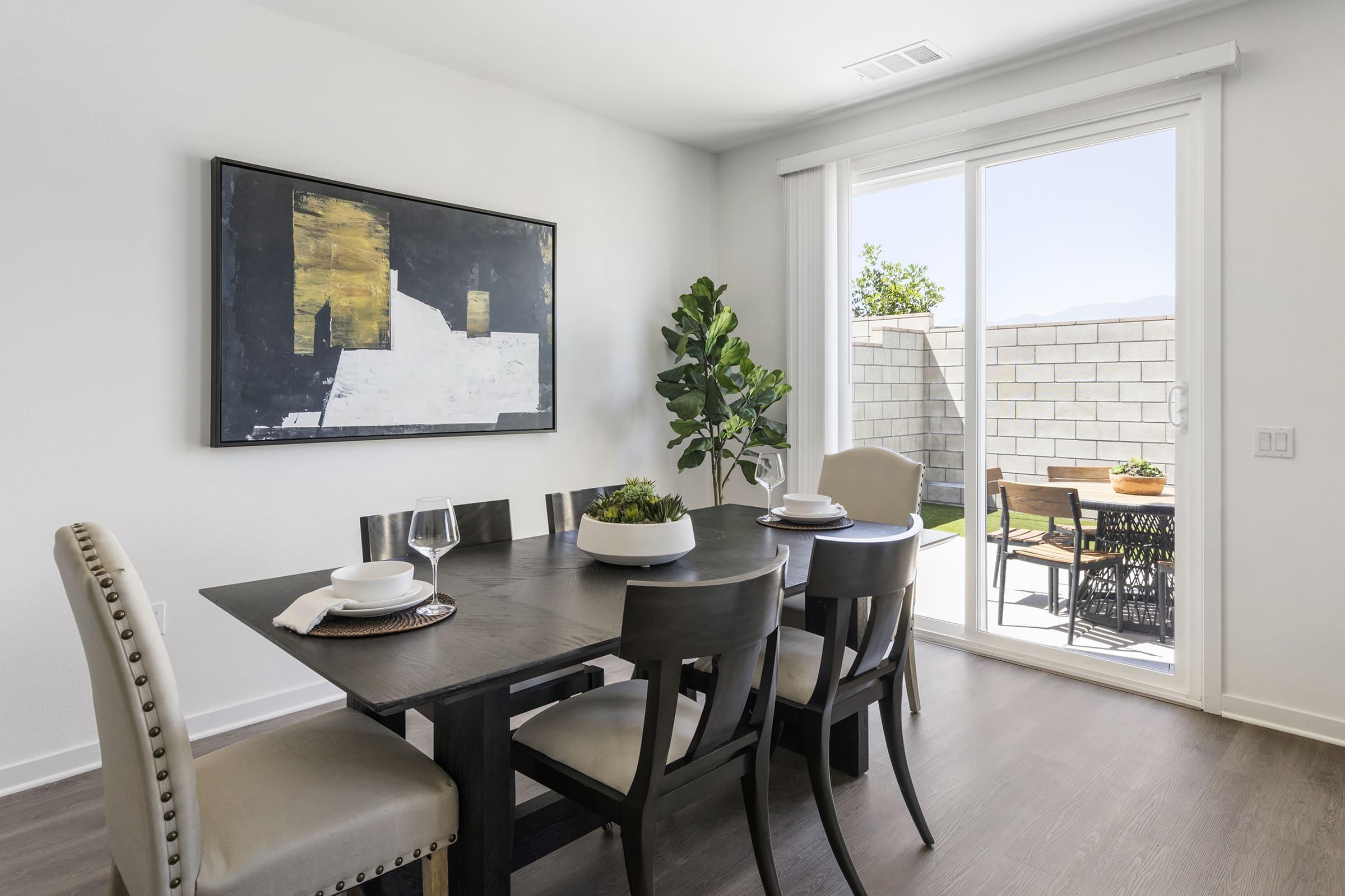 A modern dining room featuring a dark wood table set with plates and wine glasses. Soft beige chairs surround the table. A large piece of abstract art hangs on the wall. A sliding glass door leads to an outdoor patio with a small dining area and greenery visible.