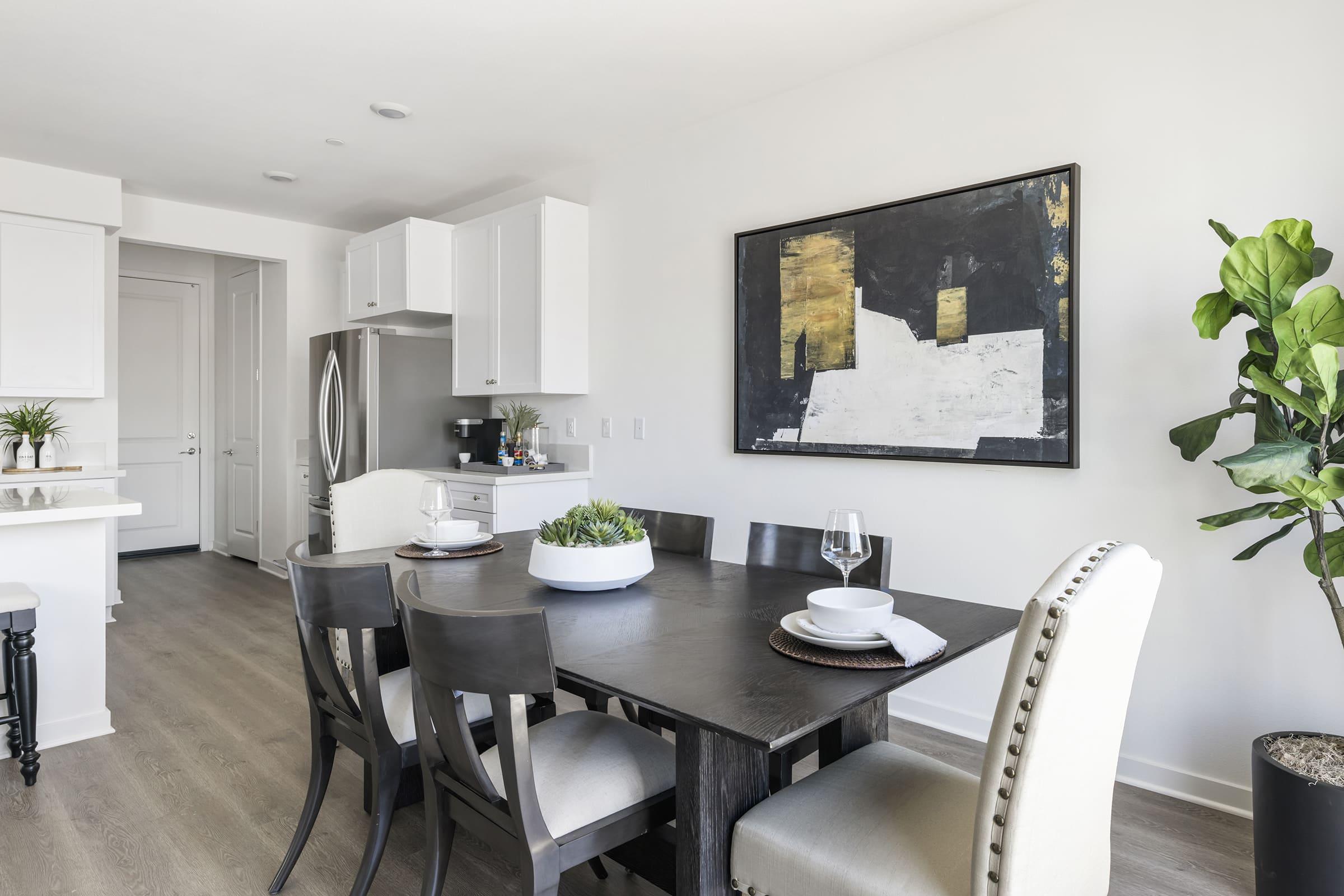 A modern dining area featuring a dark wooden table surrounded by six chairs. A large abstract painting hangs on the white wall. A potted plant adds a touch of greenery. The space is bright and minimalistic, complemented by sleek cabinetry and a stainless steel refrigerator.