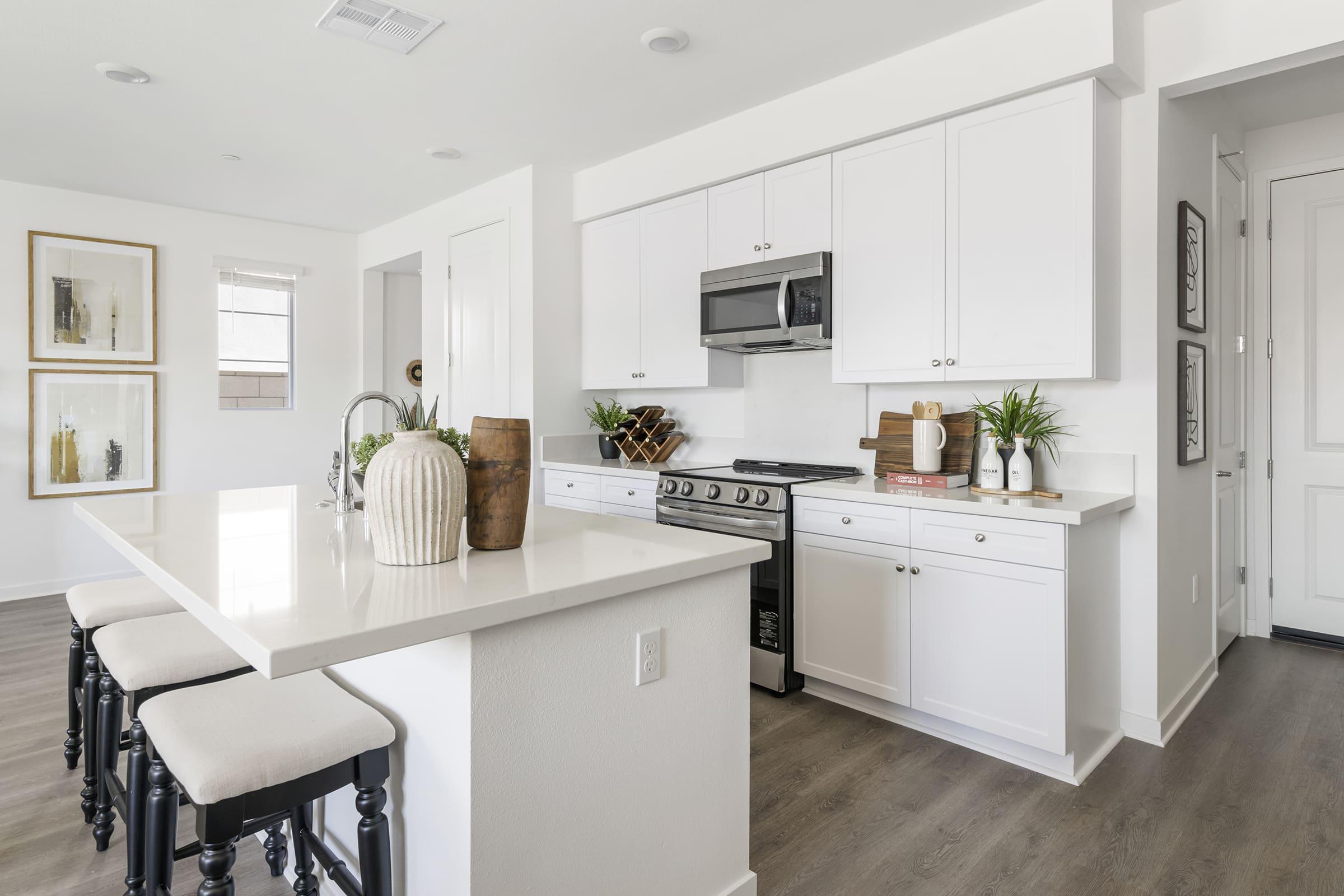 Modern kitchen featuring white cabinetry, a stainless steel microwave, and a gas stove. A large island with black barstools offers additional seating. Decorative elements include potted plants and artwork on the walls. The space is well-lit with natural light coming through the windows, contributing to a bright and inviting atmosphere.