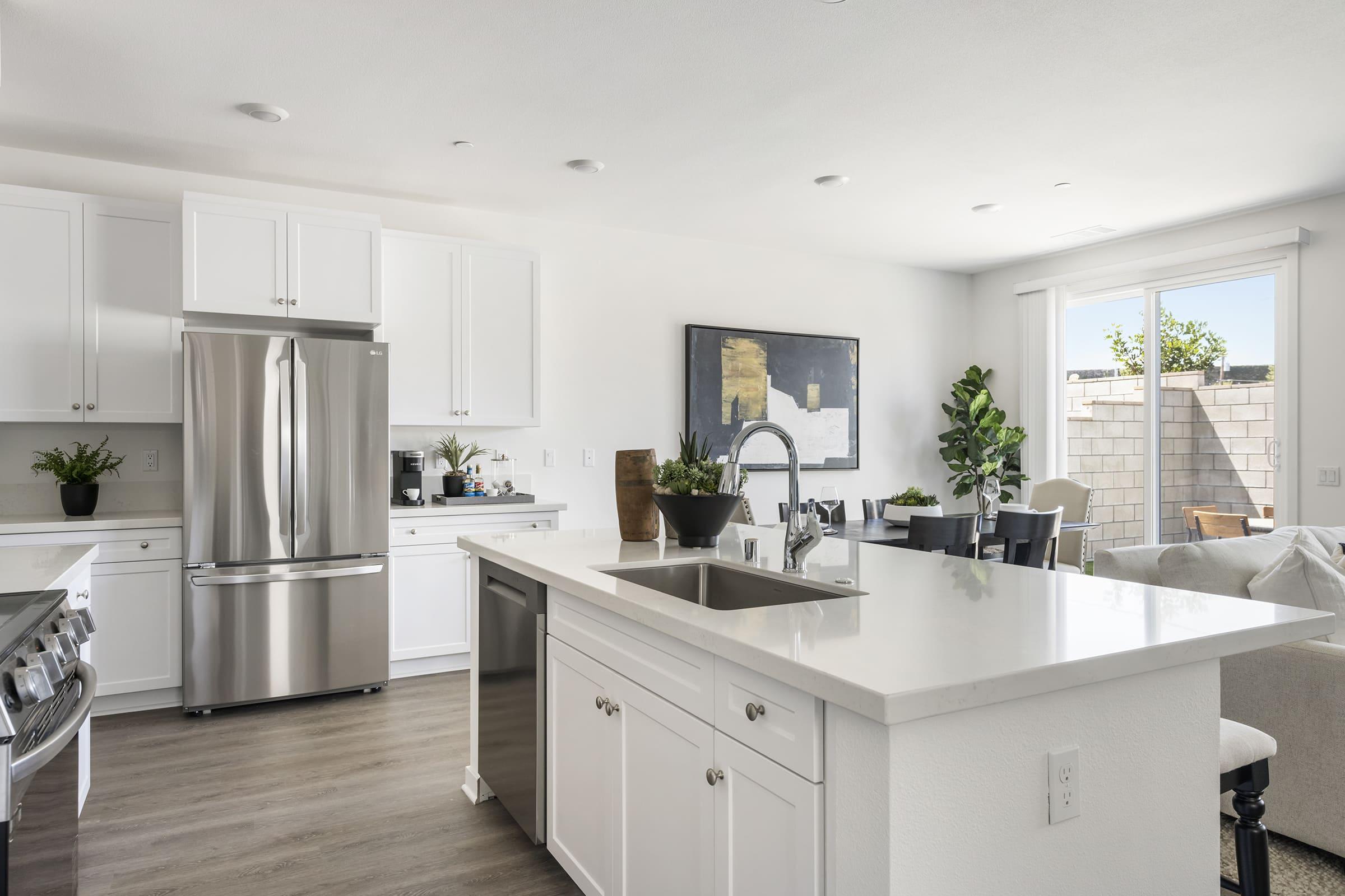 Modern kitchen featuring stainless steel appliances, white cabinetry, and a large island with a sink. The space is decorated with potted plants and has a dining area with a wooden table. Natural light floods in through large windows, creating an inviting and spacious atmosphere.